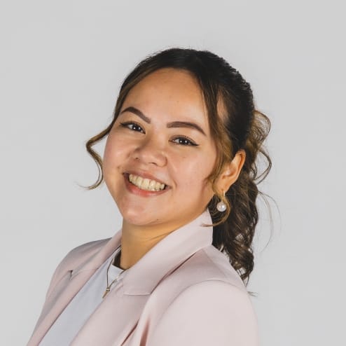 Vrouw met krullend haar in een lichtroze blazer, lachend tegen een eenvoudige achtergrond.