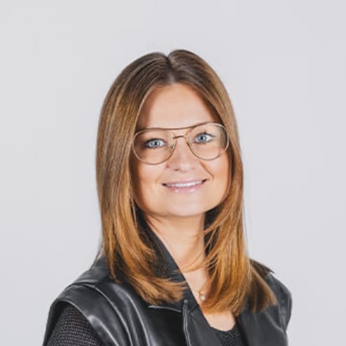 Een vrouw met een bril, stijl bruin haar, in een zwart topje tegen een lichte achtergrond, lacht.