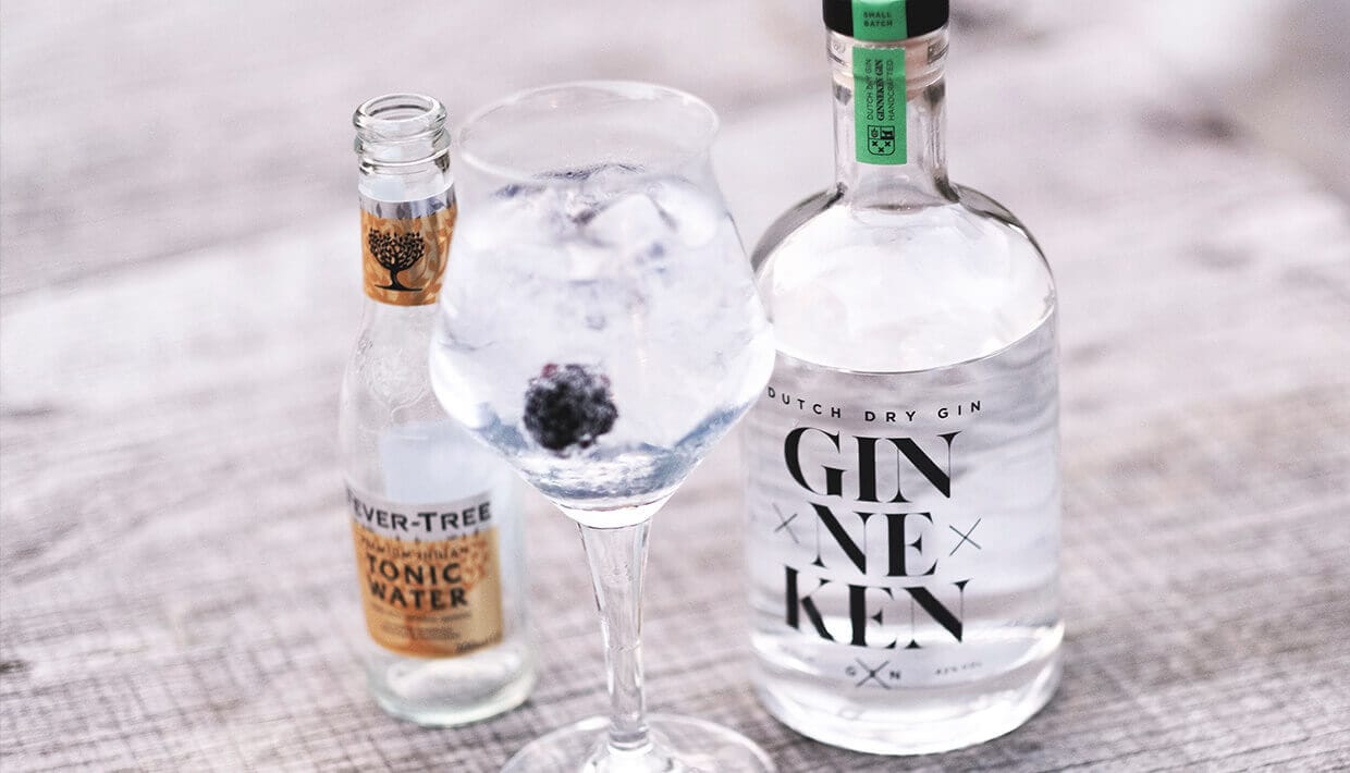 Een gin-tonic met een braambes naast flessen van GinnekenGin.com staat stijlvol op tafel.