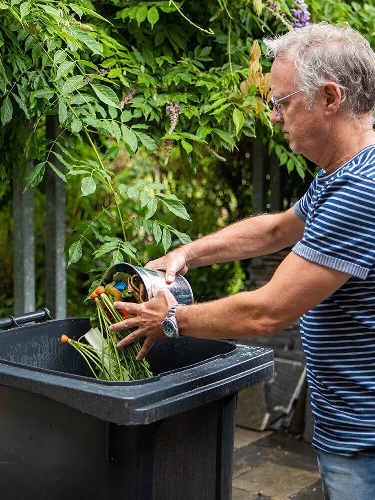 Een man in strepen gooit plantenstengels in een zwarte bak onder een bladerdak, wat de Samen Aan De Bak!-gedachte belichaamt.