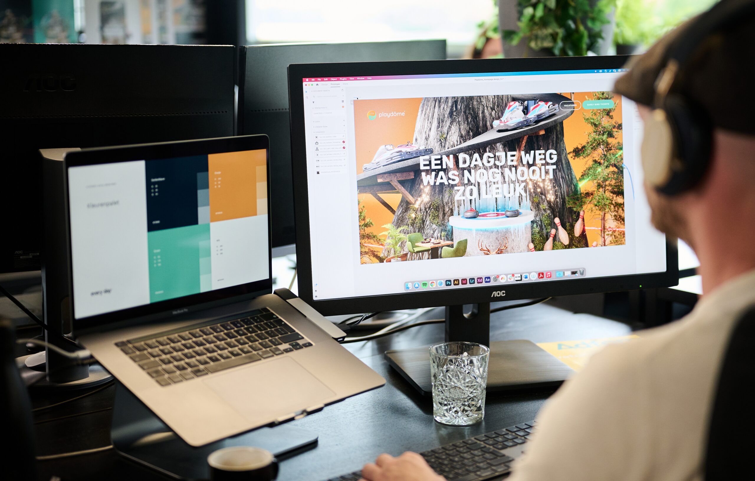 Koptelefoon op, website ontwerpen met twee monitoren en planten en water in de buurt.