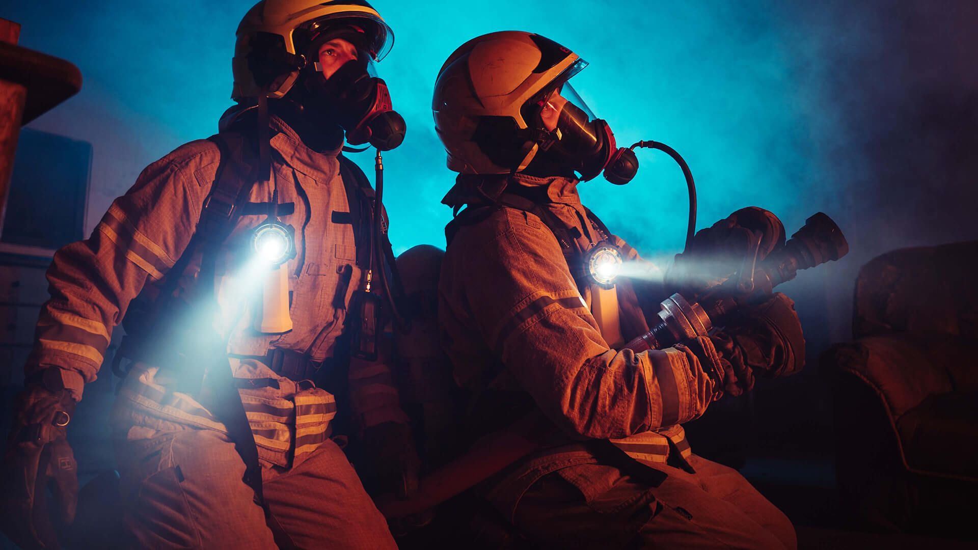 Twee gemaskerde brandweermannen in helmen gebruiken zaklampen en een slang om door een rokerige kamer te navigeren.