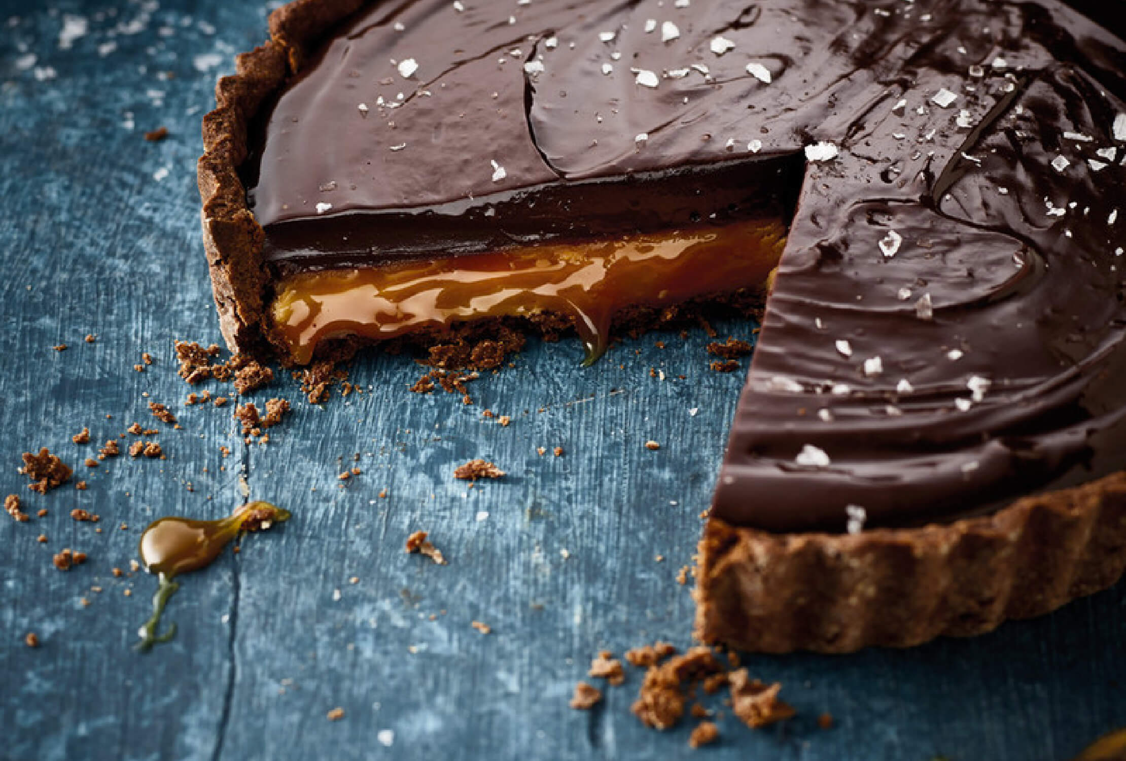 Een in punten gesneden chocolade-karameltaartje, waarvan het smeuïge midden zichtbaar is, bedekt met zeezoutvlokken op een blauw oppervlak.