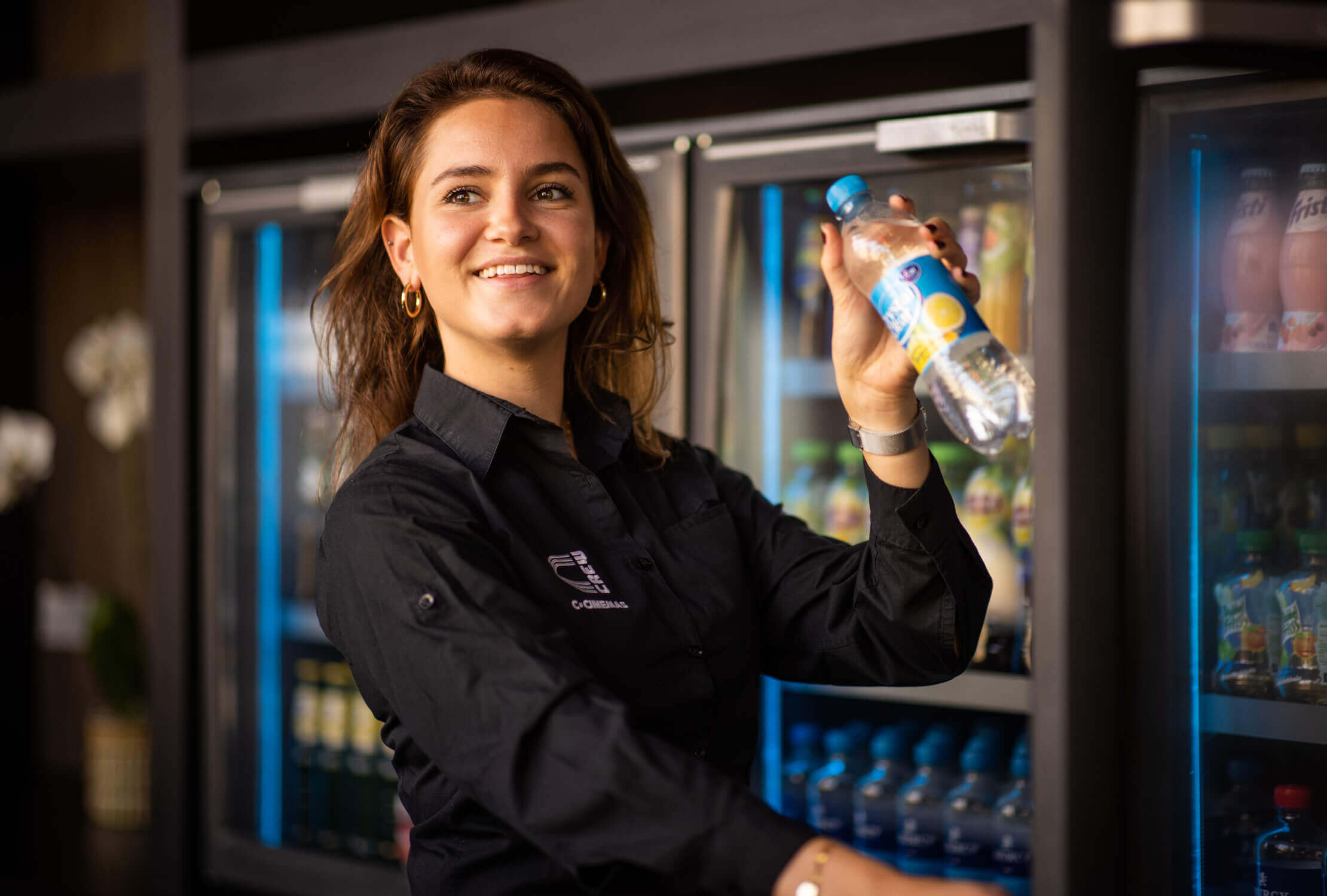 Vrouw in zwart shirt lacht met fles in haar hand bij de koelkast.