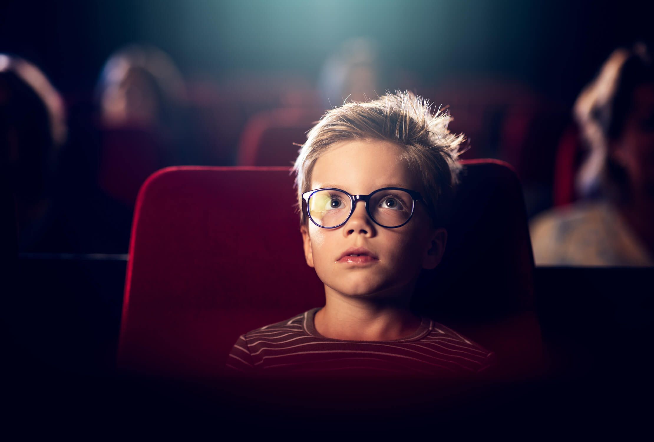 Een jongen met een bril kijkt in een schemerige bioscoopzaal naar een film, zittend op rode bekleding.