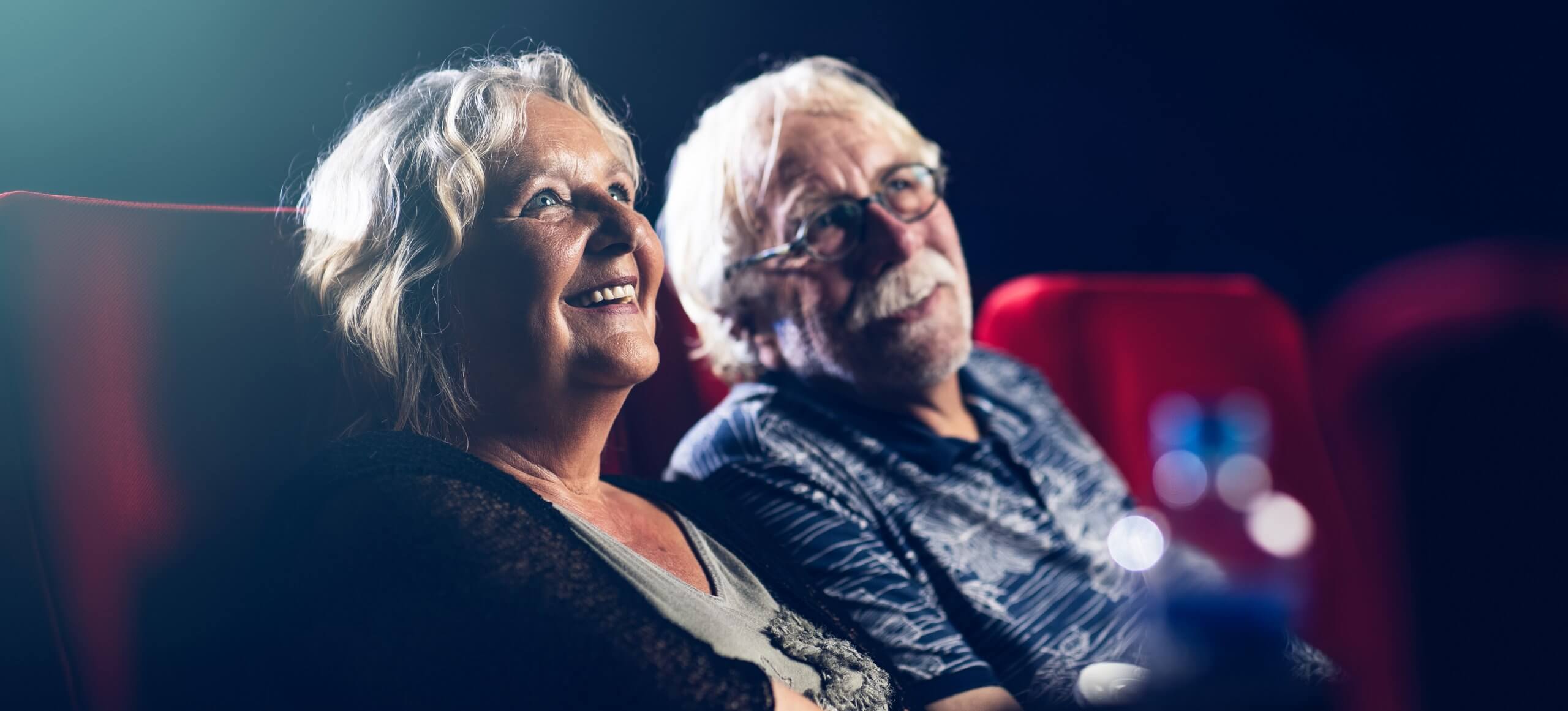Een ouder echtpaar lacht terwijl ze genieten van een film in een theater met rode stoelen.
