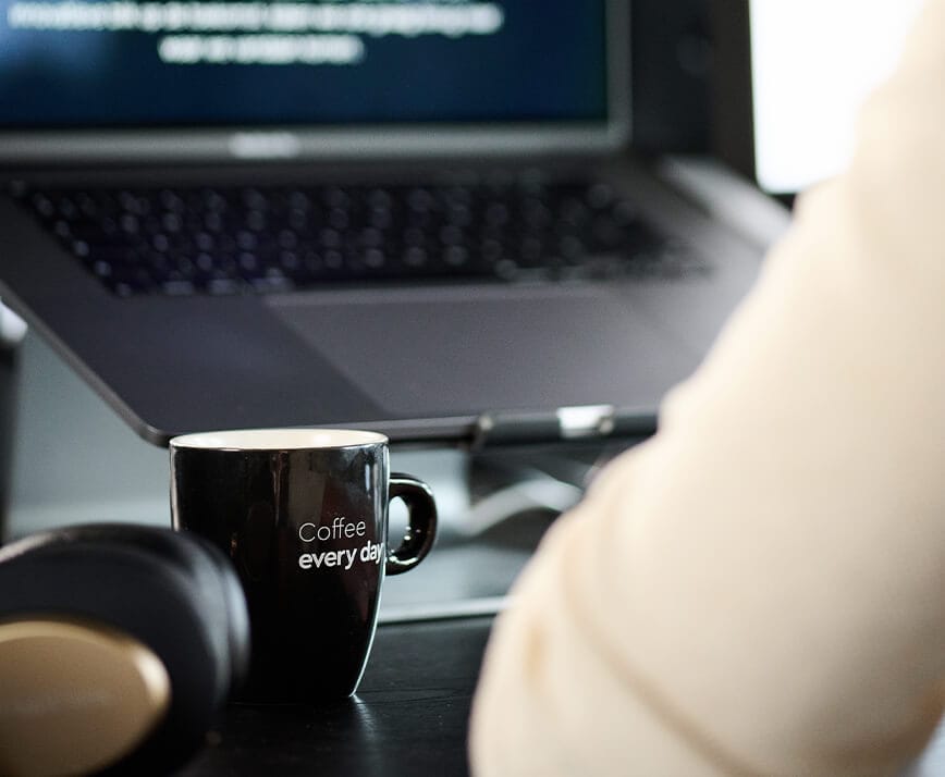 Een zwarte mok met de tekst "Koffie voor elke dag" staat op een bureau, naast een laptop en een koptelefoon.