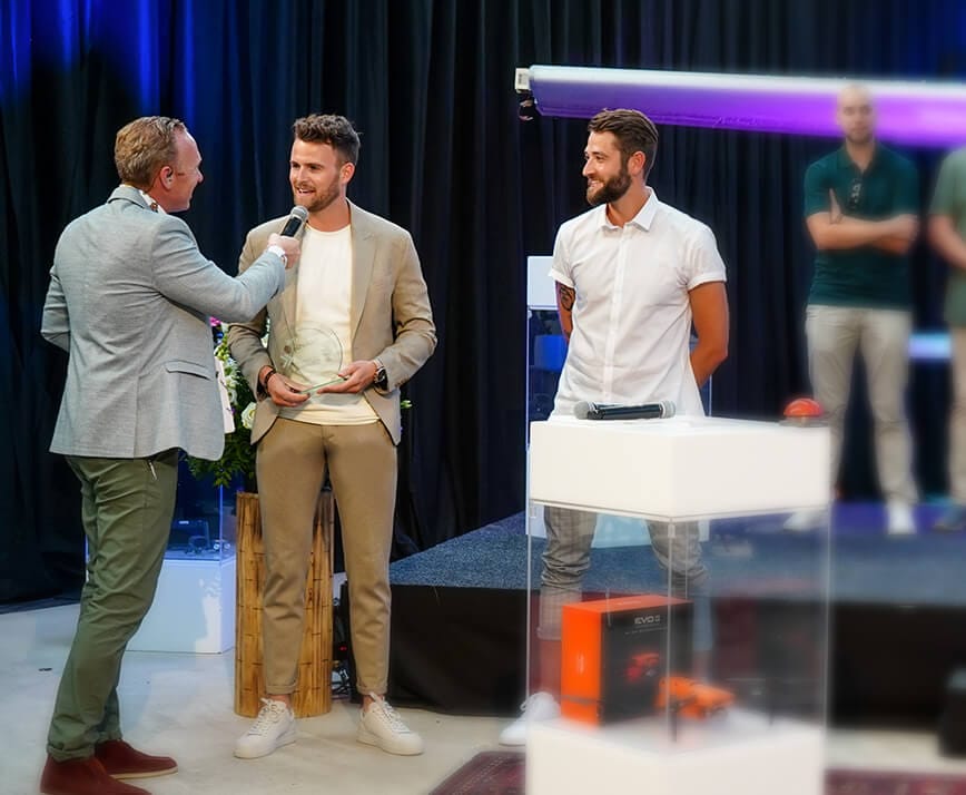 Twee mannen staan, één met een microfoon, met een wazig publiek op de achtergrond.