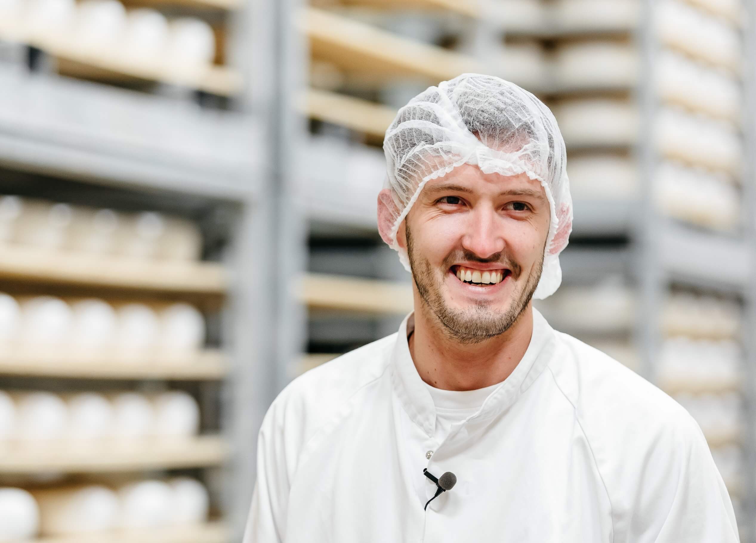 Lachende man in een wit pak en haarnetje staat in een opslagruimte met planken vol ronde voorwerpen.
