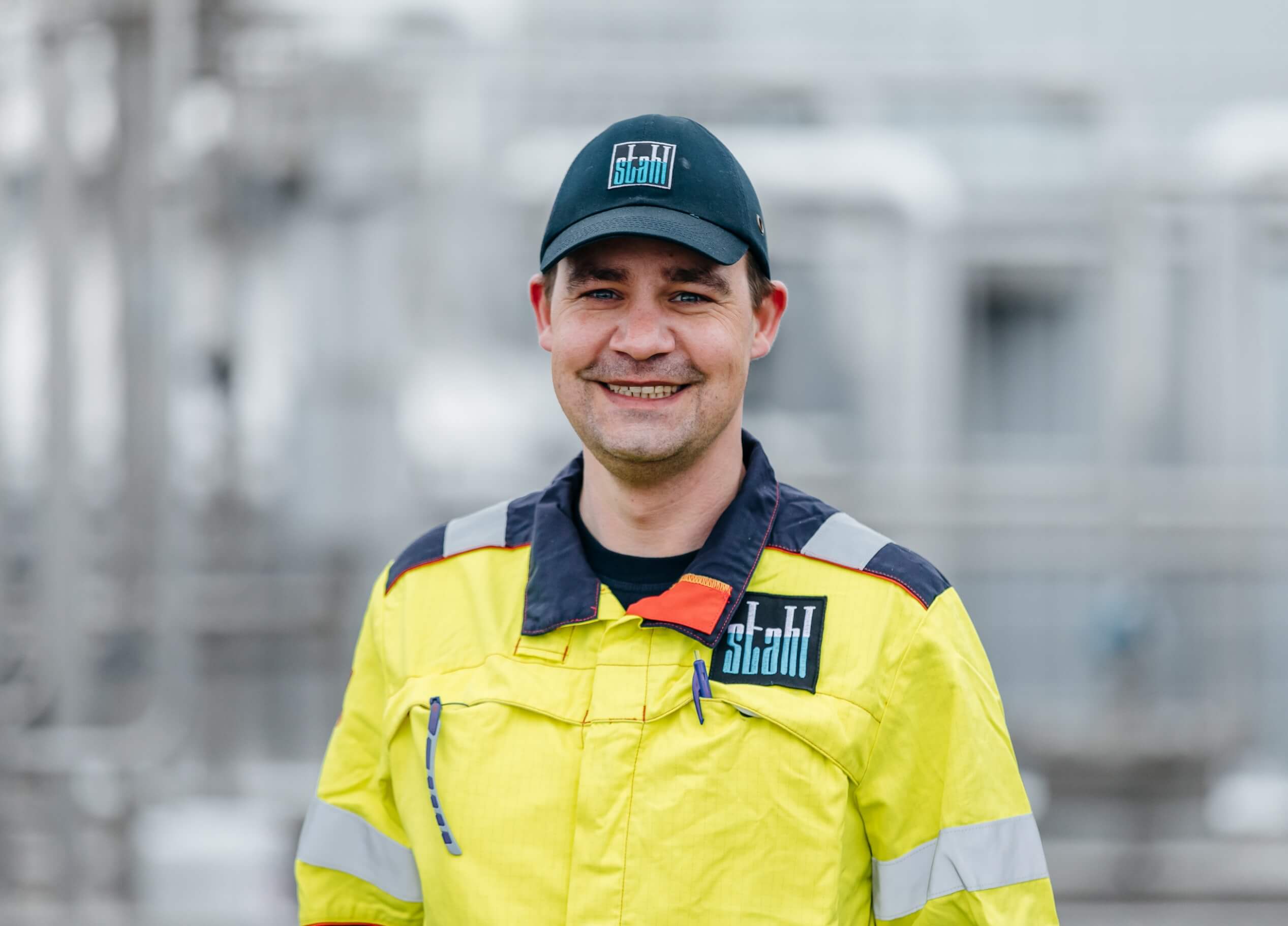 Een man lacht in een geel uniform en een zwarte Stahl-pet, tegen een industriële achtergrond.