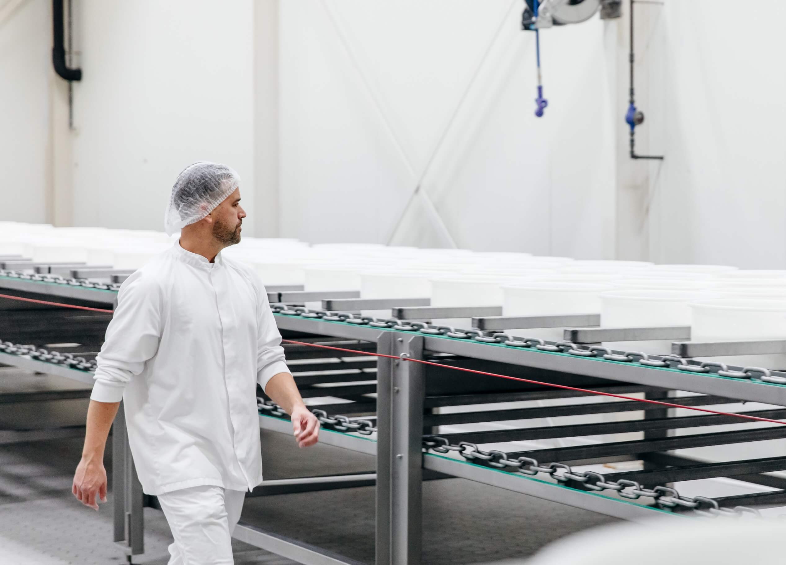 Een fabrieksarbeider in een wit uniform en met een haarnetje loopt langs transportbanden in een industriële omgeving.