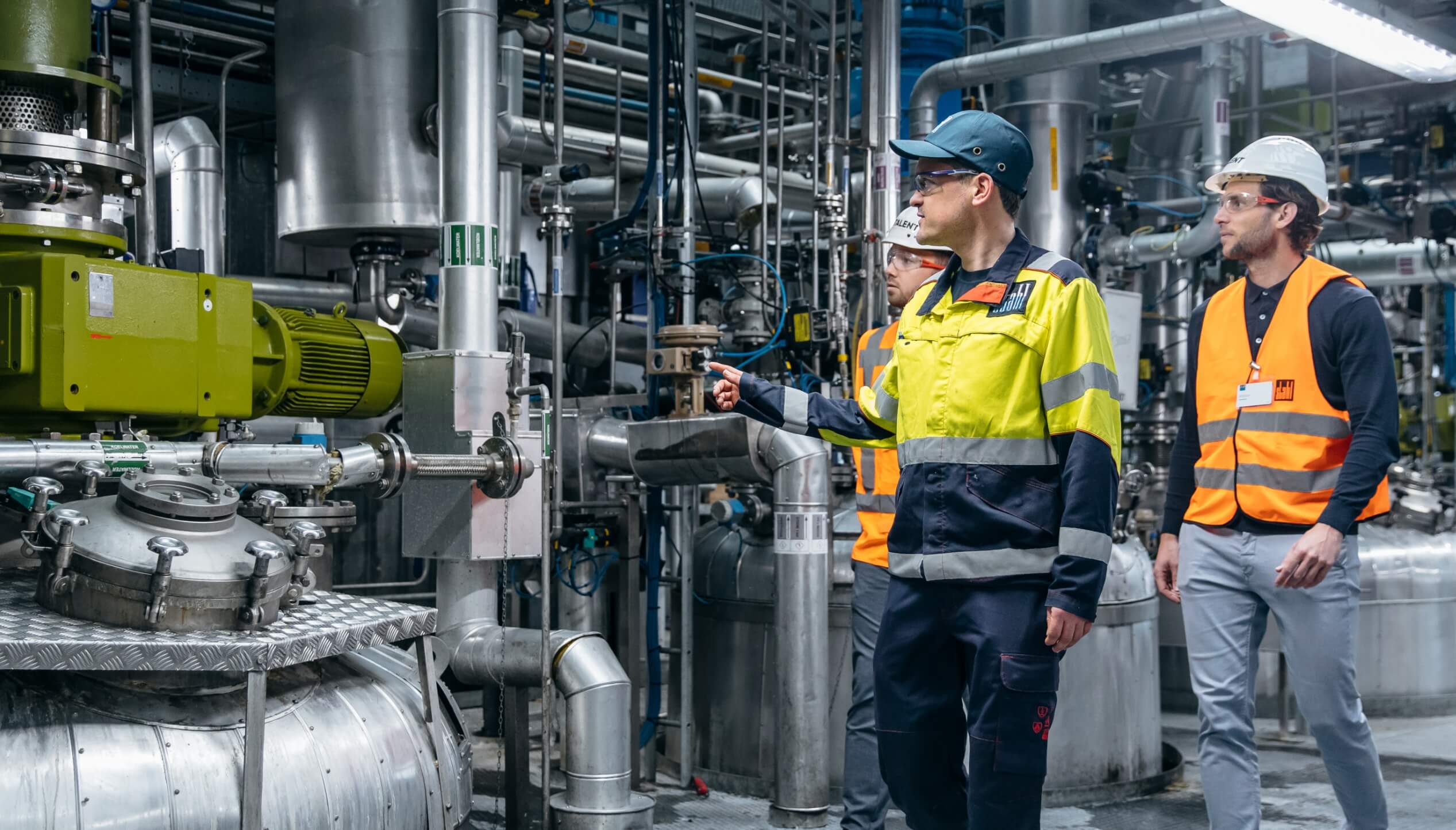 Drie werknemers in veiligheidskleding inspecteren de machines in een fabriek.