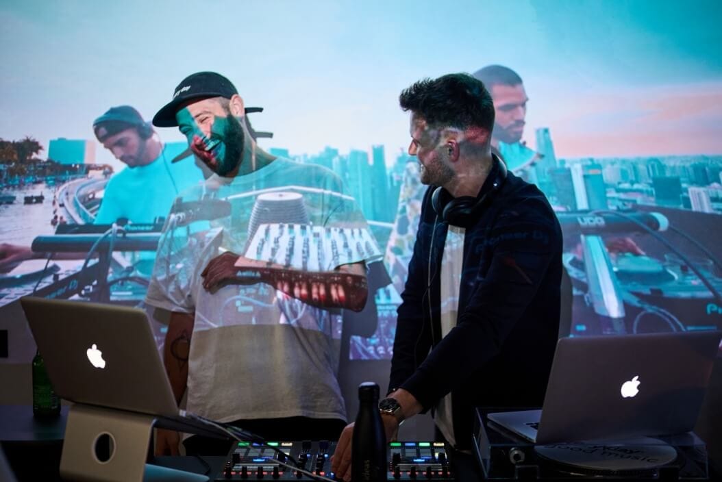 Twee DJ's dansen met laptops en mixers, terwijl de skyline van de stad op hen wordt verlicht.