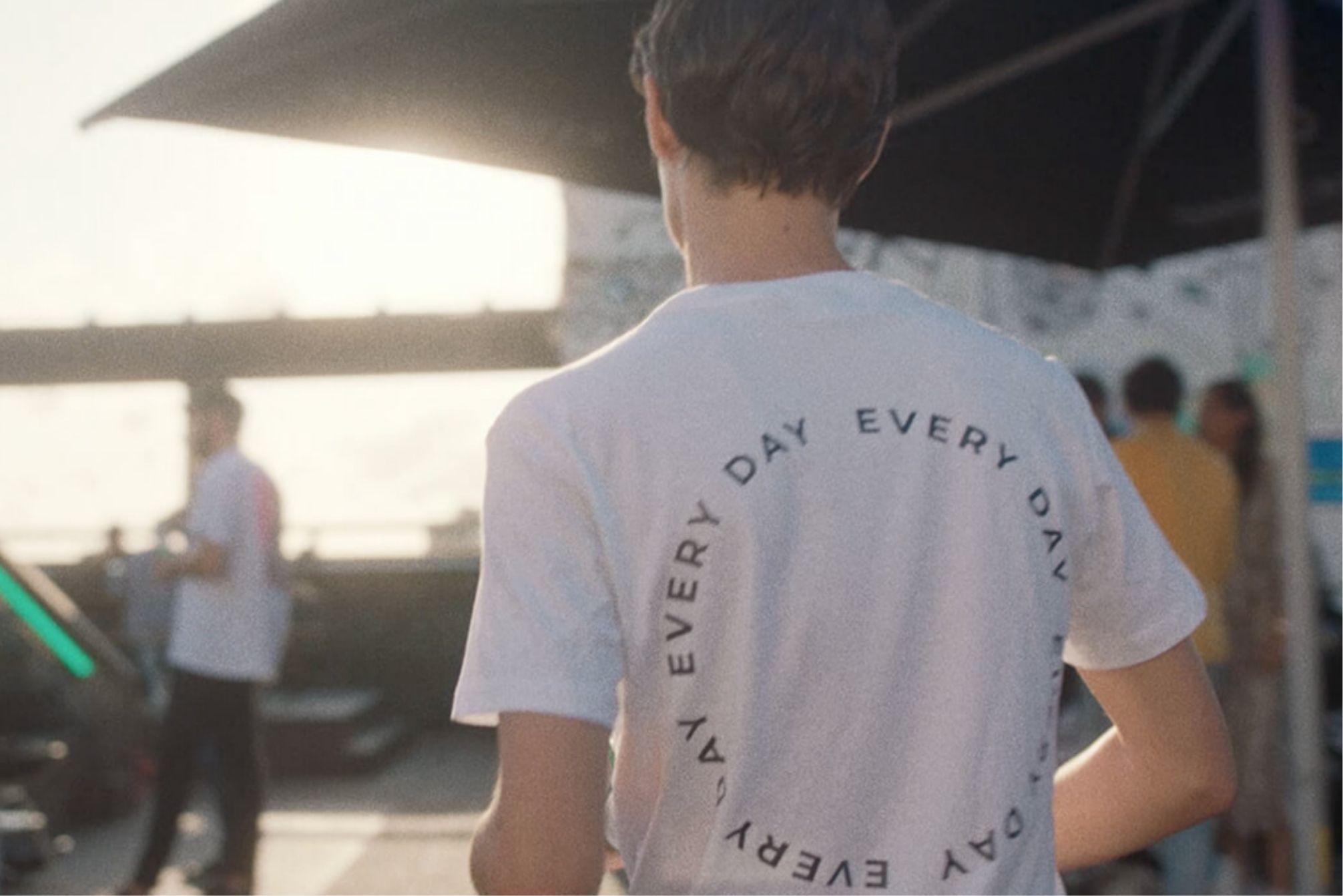 Persoon in 'EVERY DAY'-shirt staat buiten; op de achtergrond zijn wazige mensen en paraplu's te zien.