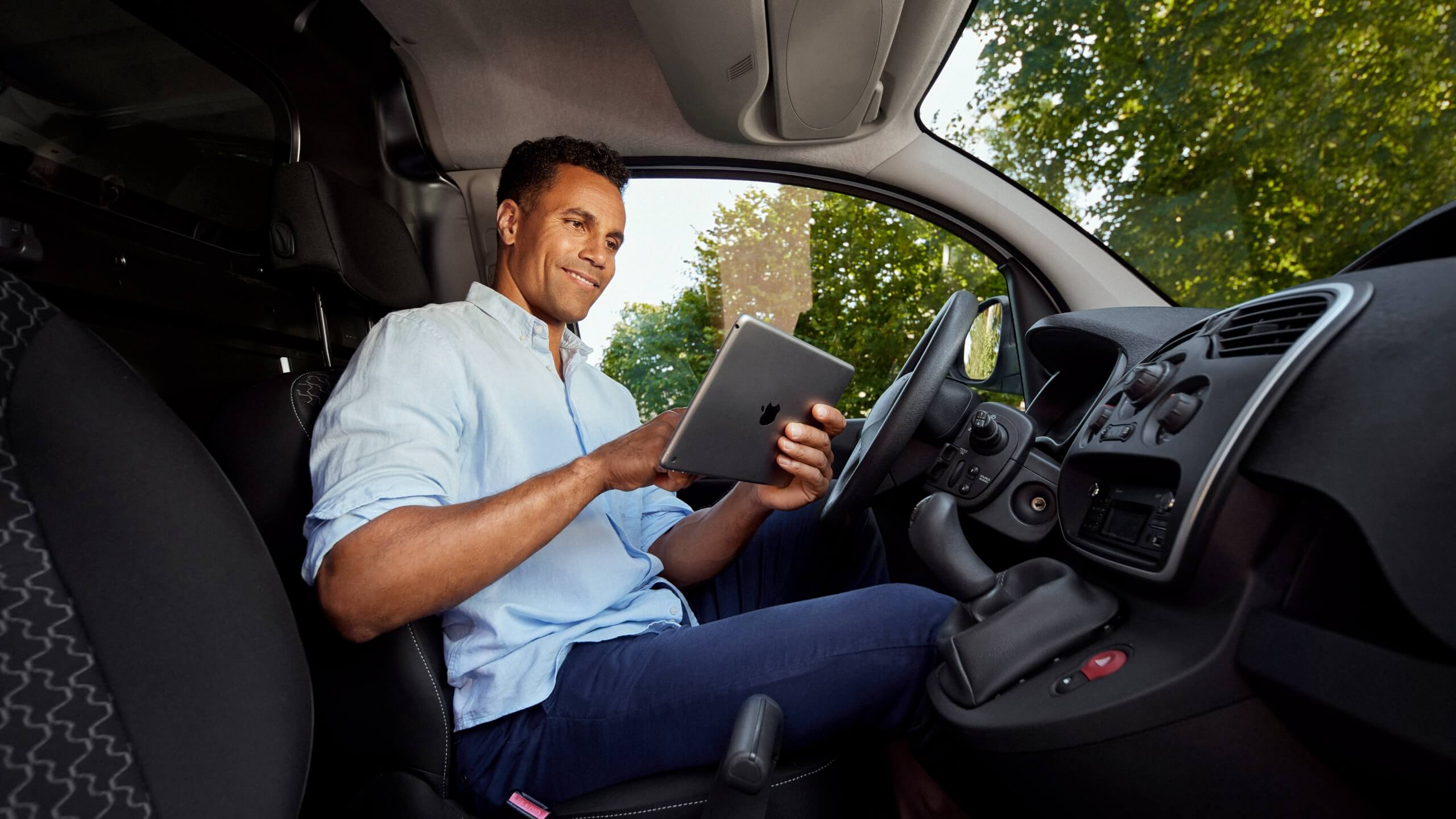 Man in een lichtblauw shirt die een tablet gebruikt in een geparkeerde auto, met bomen zichtbaar door het raam.