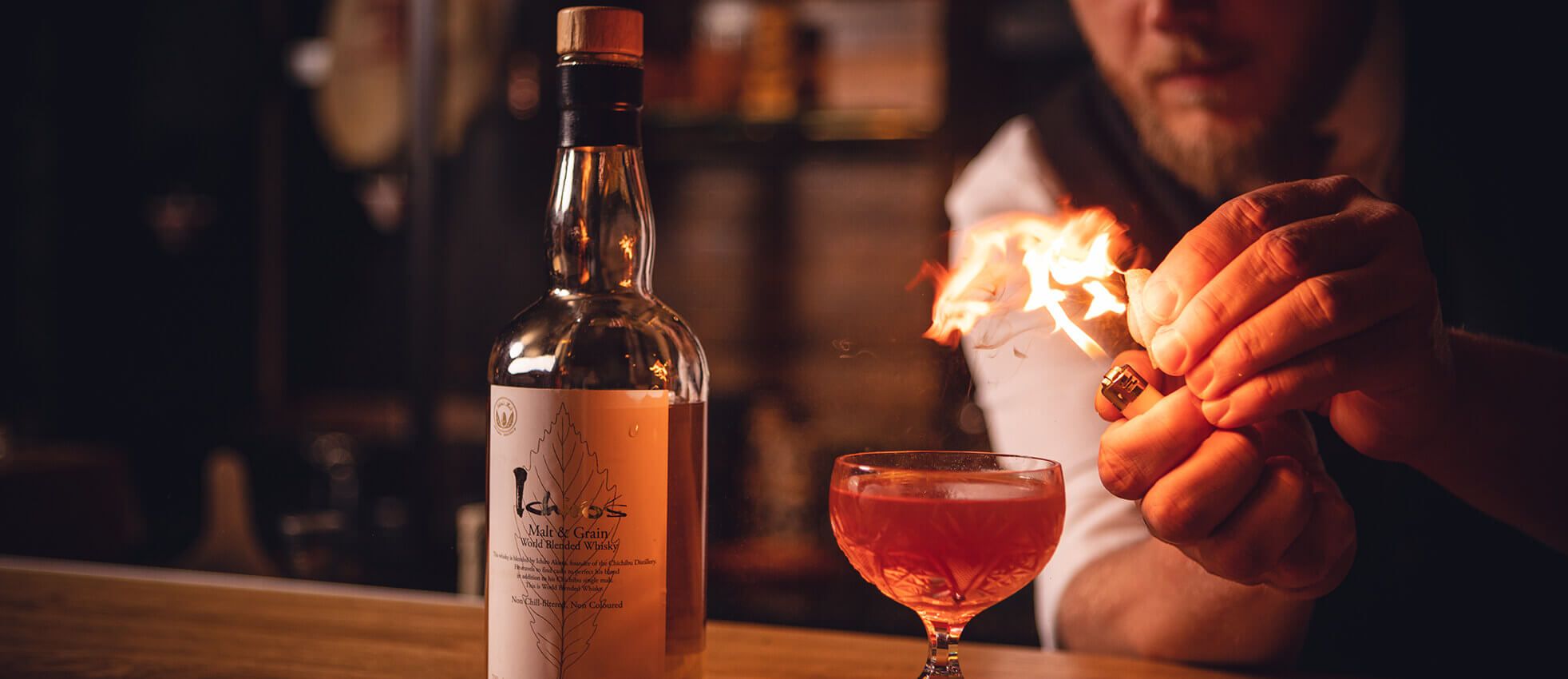 Een barman steekt een cocktail aan naast een fles op de houten bar.
