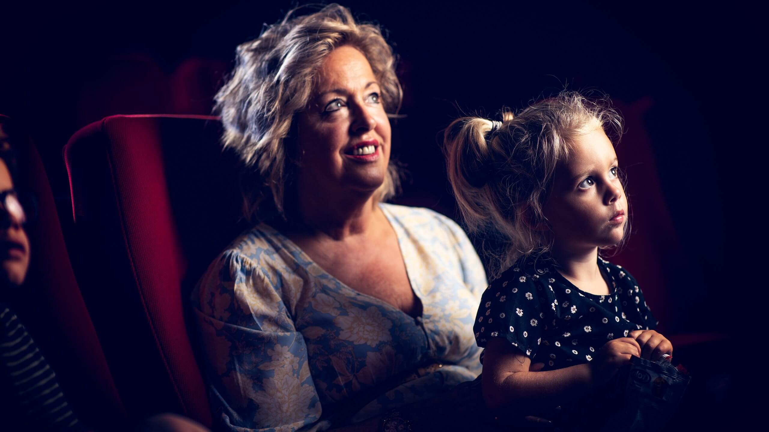 Een vrouw en een kind zitten aandachtig in een donker theater, verzonken in de voorstelling.