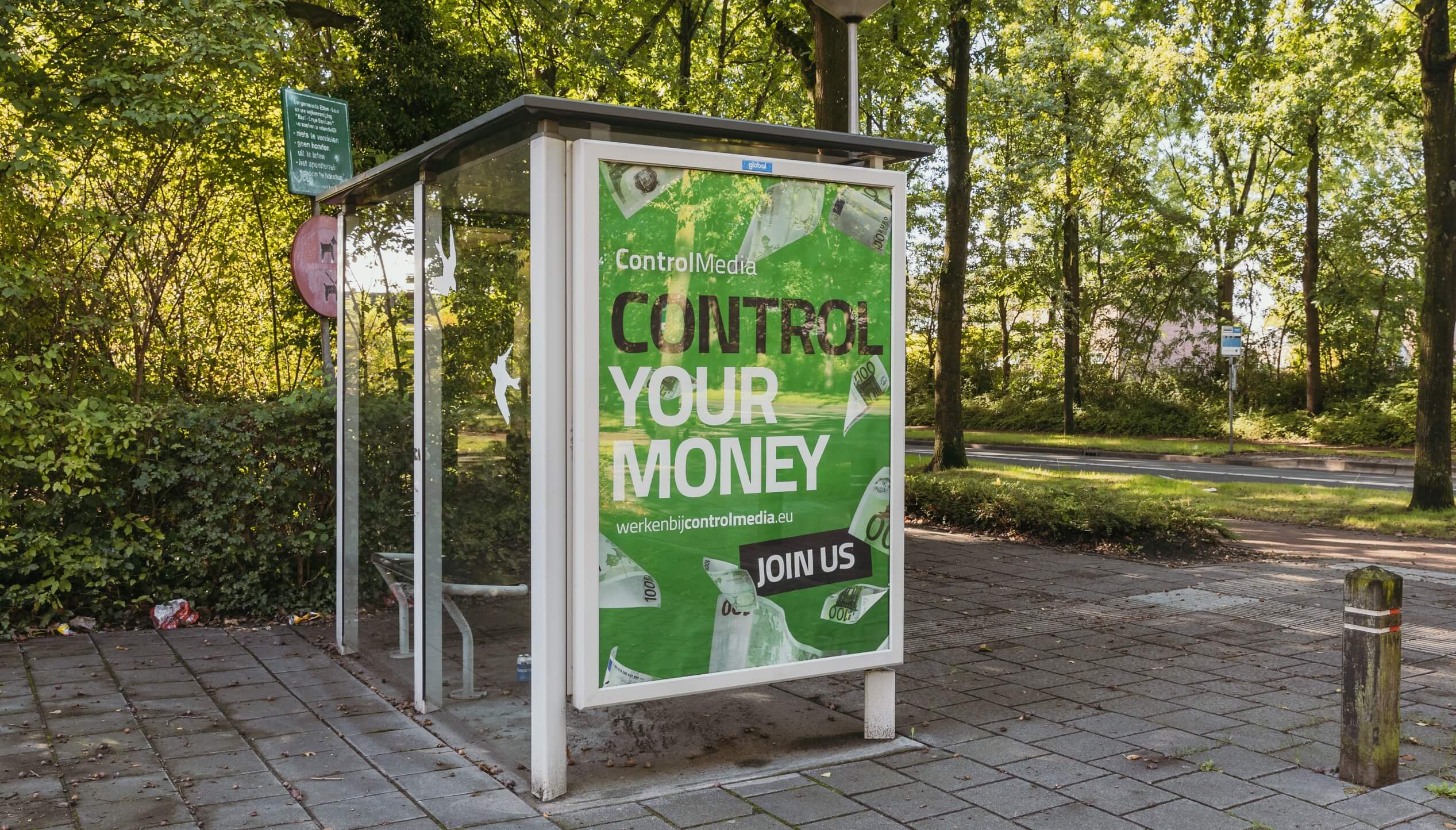 Advertentie bij bushalte: "Controleer uw geld" - groene poster, pad met bomen erlangs, naast geplaveid wandelpad.