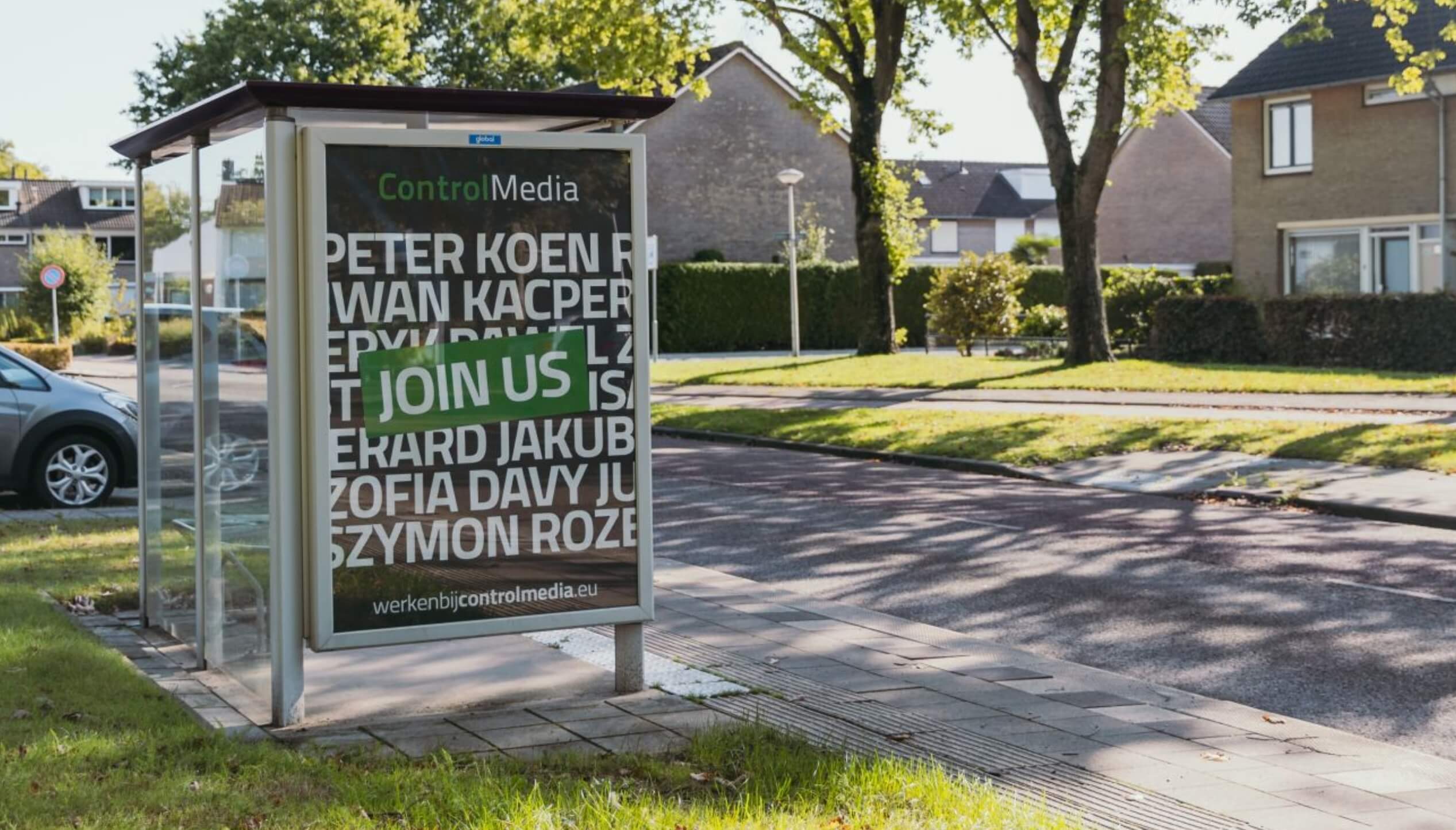 Poster bij bushalte: "Word lid van Control Media!" Woonstraat en huizen op de achtergrond.
