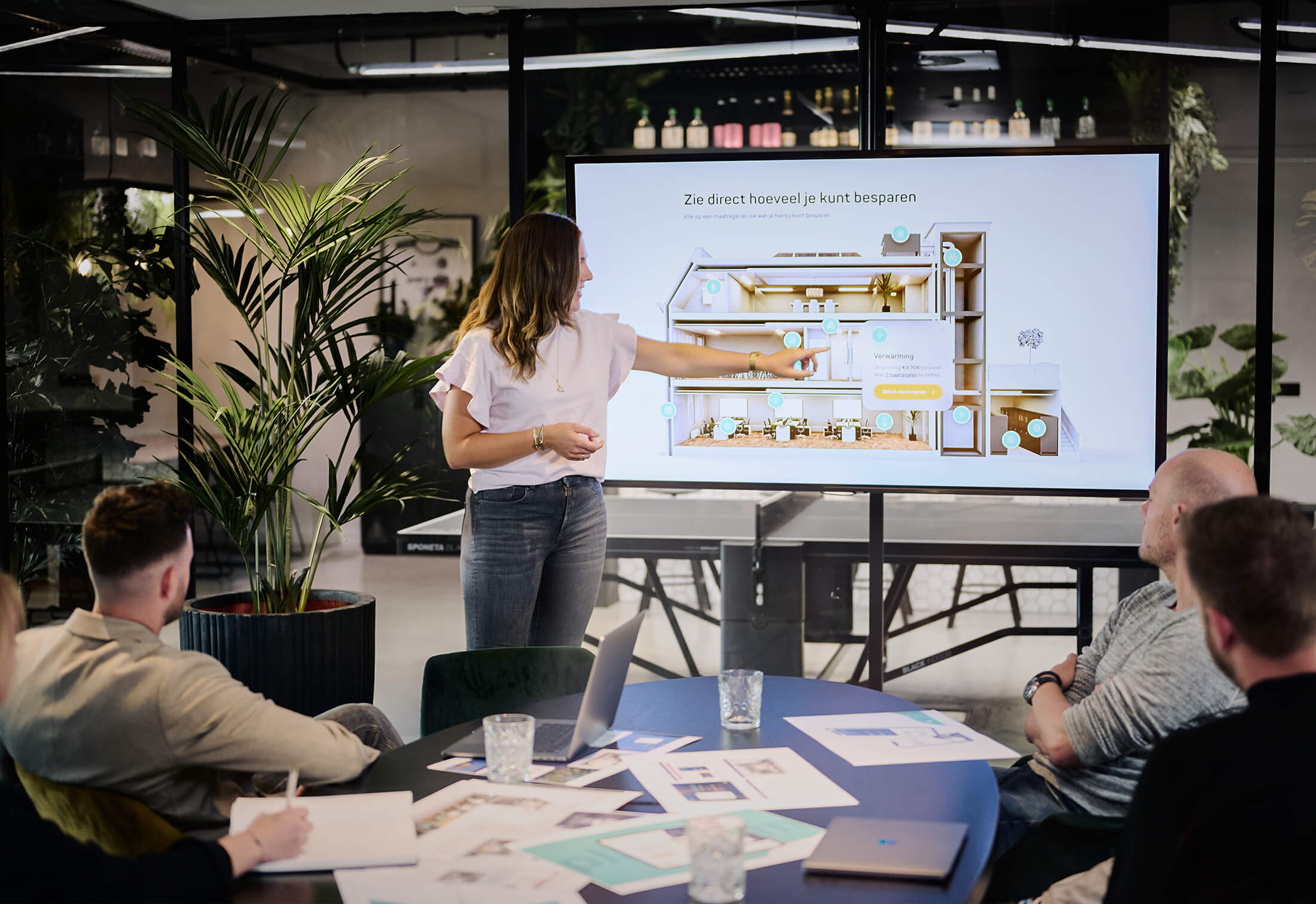 Een vrouw laat op het scherm een plattegrond zien aan drie collega's in een modern kantoor.