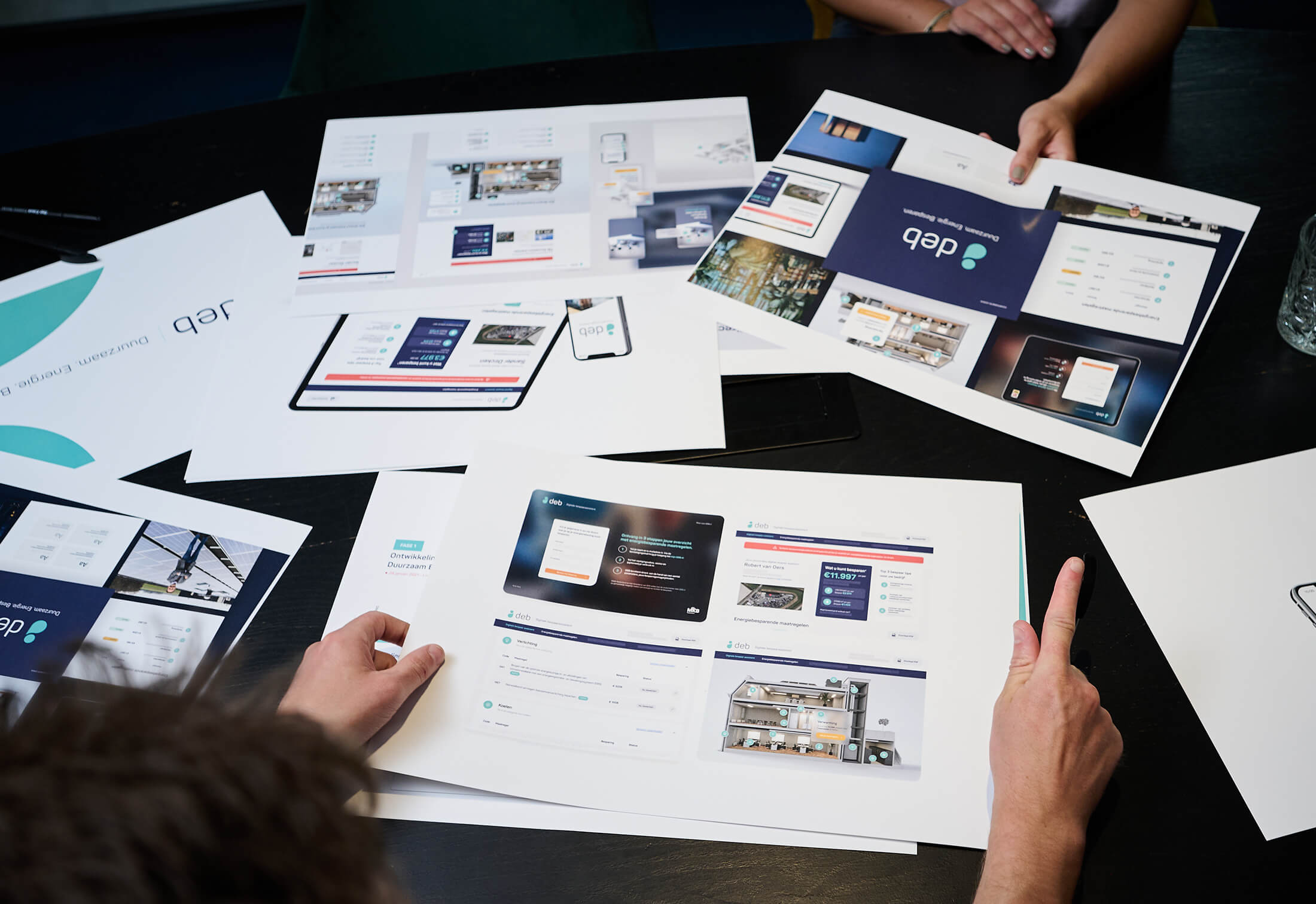 Mensen verzamelen zich rond een tafel en bekijken ontwerpmodellen op papier en apparaten.