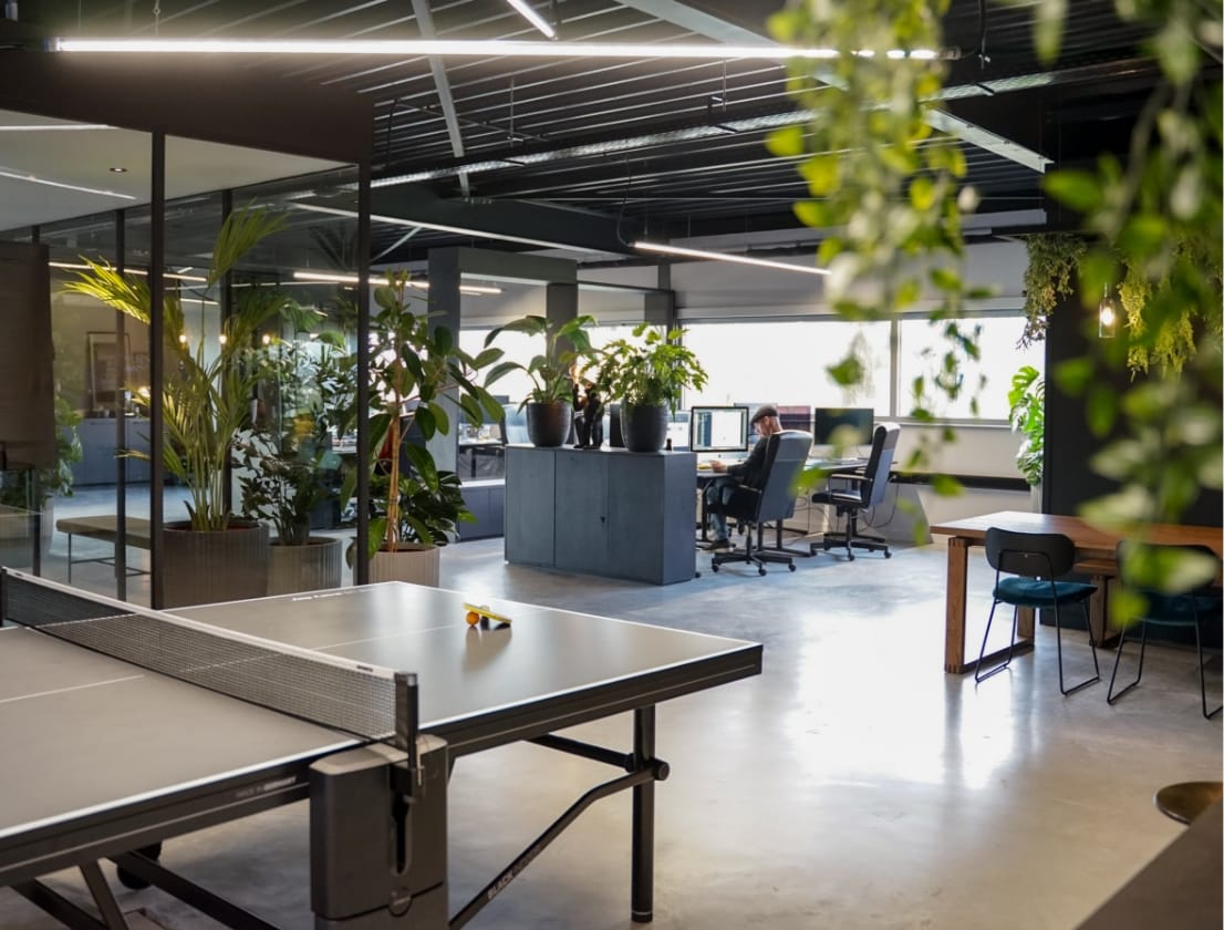Modern kantoor: bureaus met computers, tafeltennistafels, planten, alles badend in natuurlijk licht.