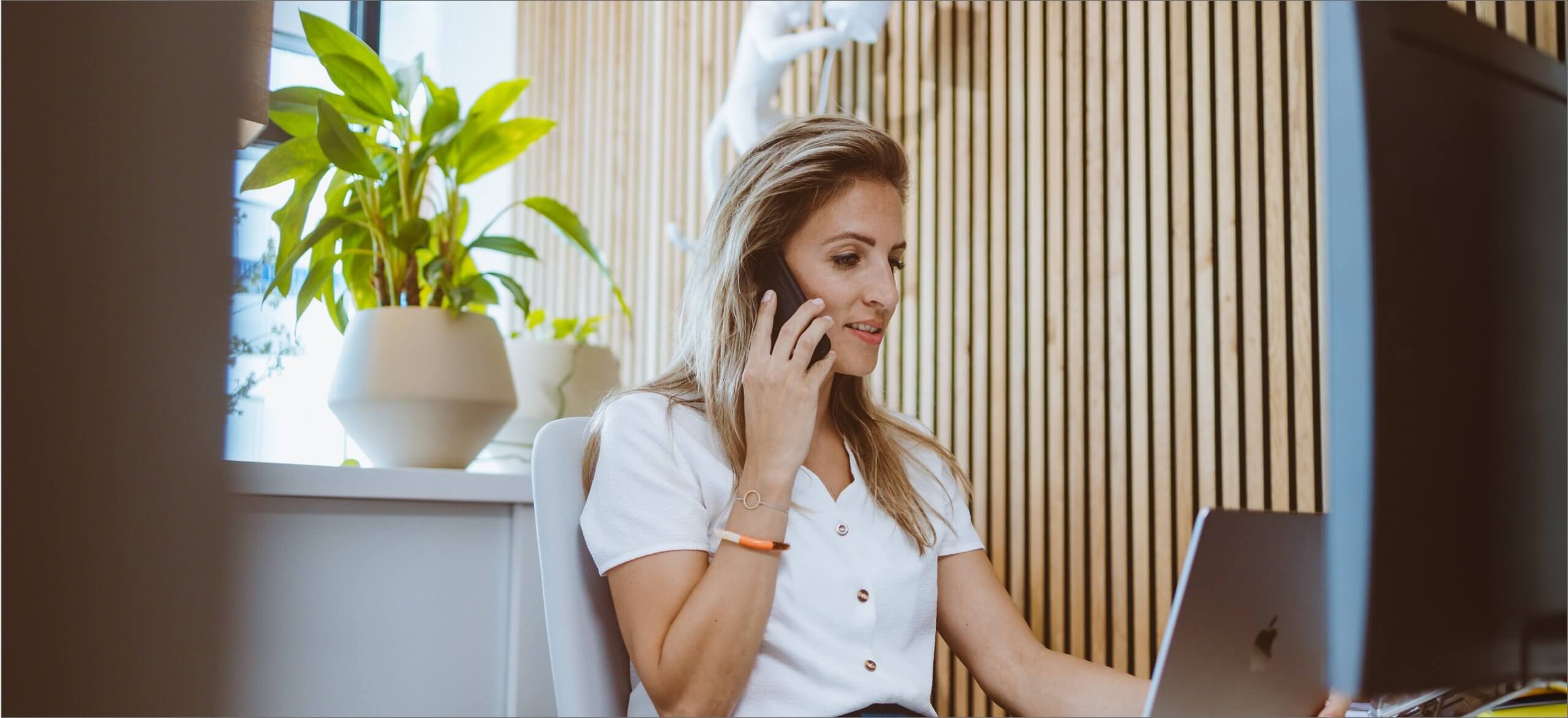 Een vrouw is aan het multitasken: ze kletst aan de telefoon en werkt op een laptop, met een potplant naast zich.