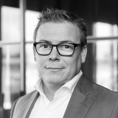 Zwart-witfoto: lachende persoon met bril en pak, binnenshuis met onscherpe achtergrond.