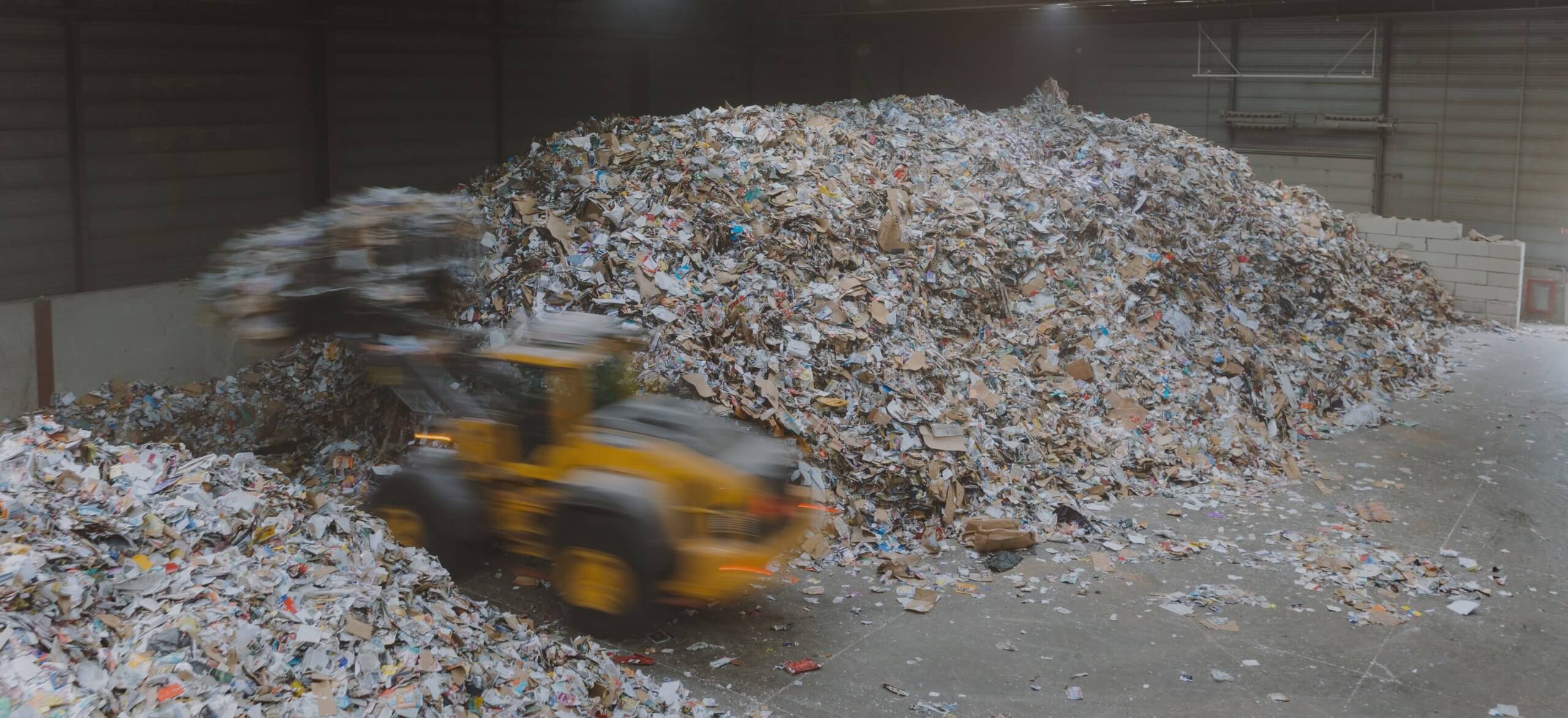 Een gele lader verplaatst papier en karton in een grote overdekte recyclingfaciliteit.