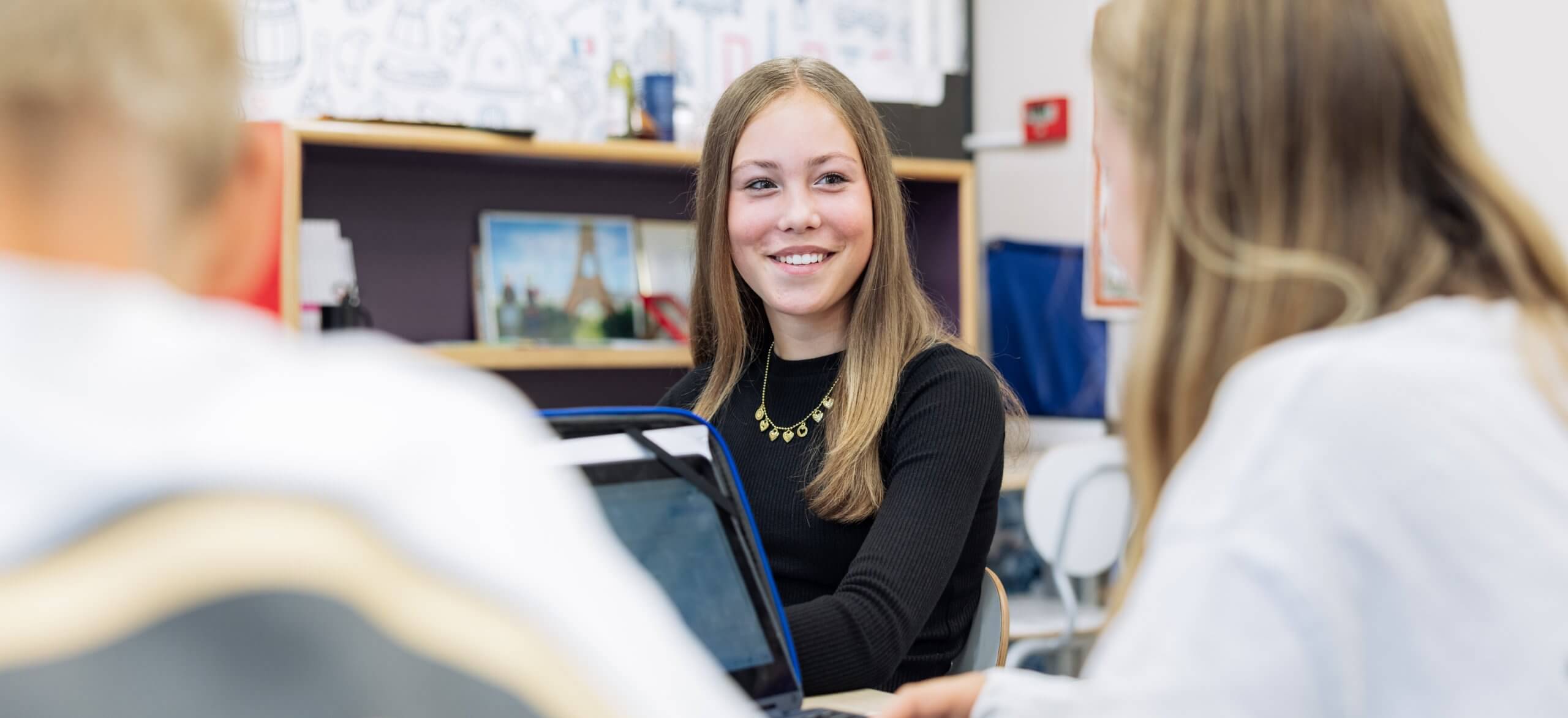 Drie studenten zitten in de klas aan een tafel te kletsen; één student zit achter een laptop.