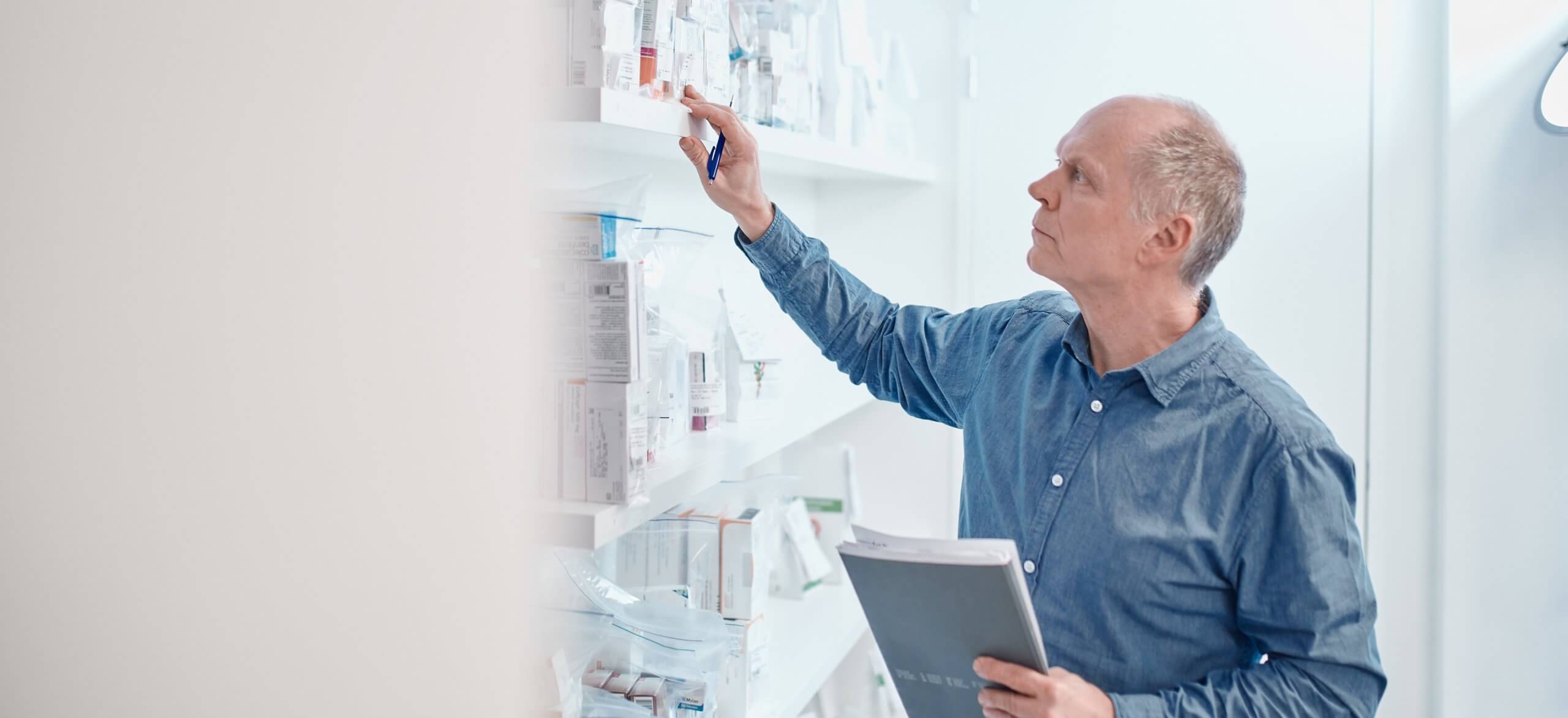 Een man in een blauw shirt pakt spullen van een plank en houdt een groot notitieboekje vast.