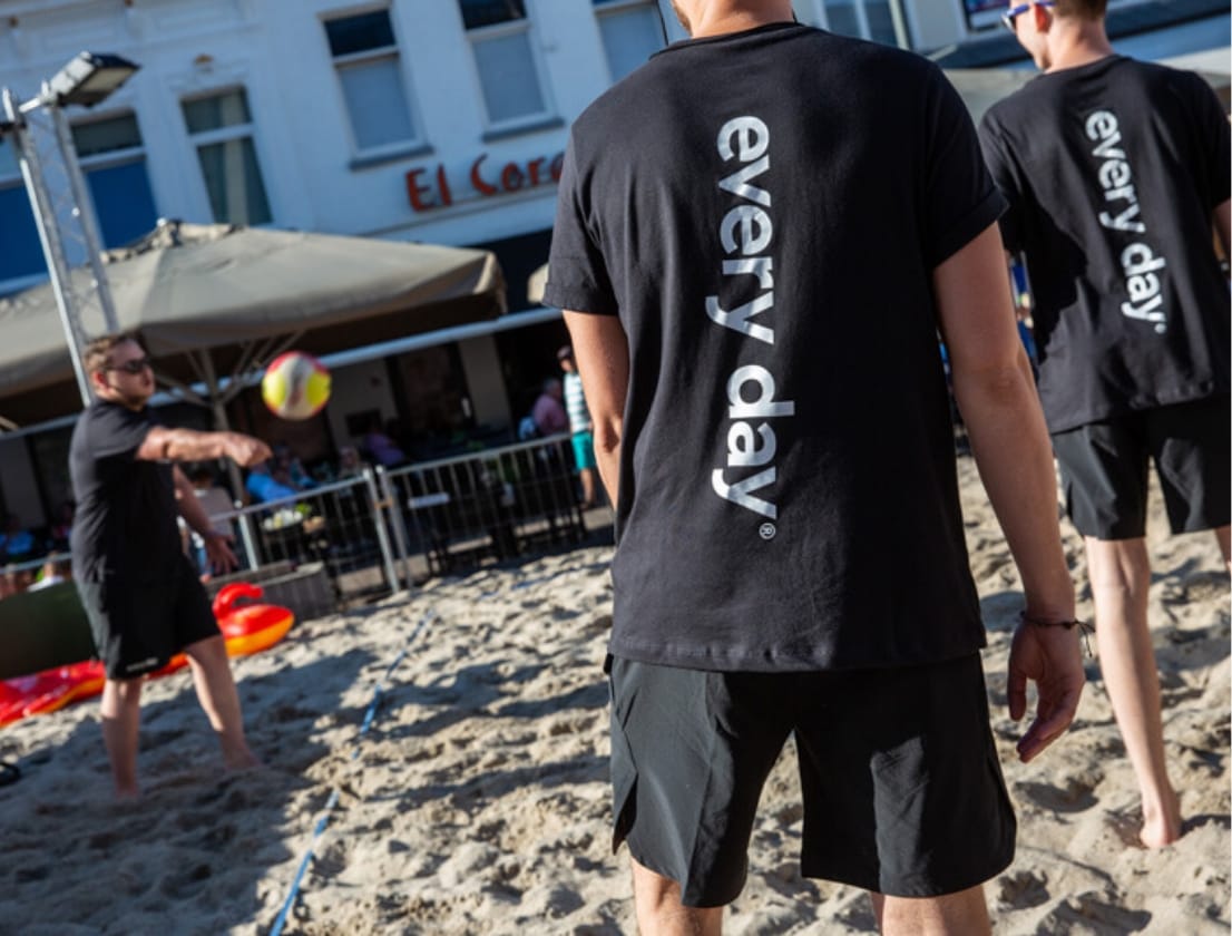 Spelers in zwarte T-shirts spelen volleybal op een zanderig terrein bij een gebouw.