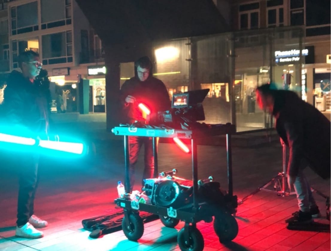 Drie personen filmen met neonverlichting in de nachtelijke stad; camera en apparatuurwagen zijn zichtbaar.