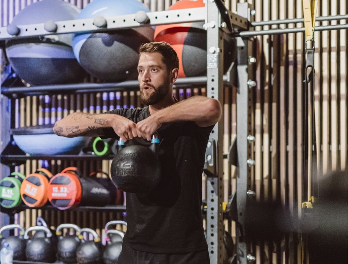 Man tilt kettlebell op in de sportschool, omringd door fitnessapparatuur en felgekleurde medicijnballen.