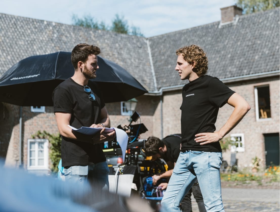 Twee mensen zijn aan het kletsen op een filmset bij een bakstenen gebouw, terwijl de camera en de crew in de buurt druk bezig zijn.