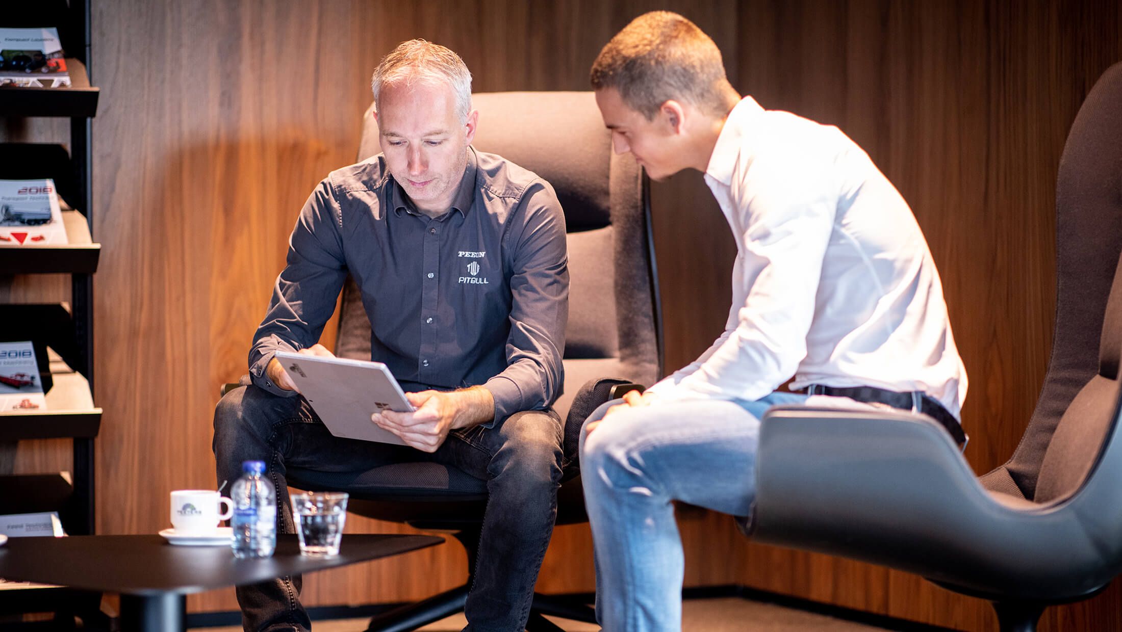 Twee mensen kletsen met elkaar op stoelen met het logo van Peeters, met een beker en een fles op een tafeltje ernaast.