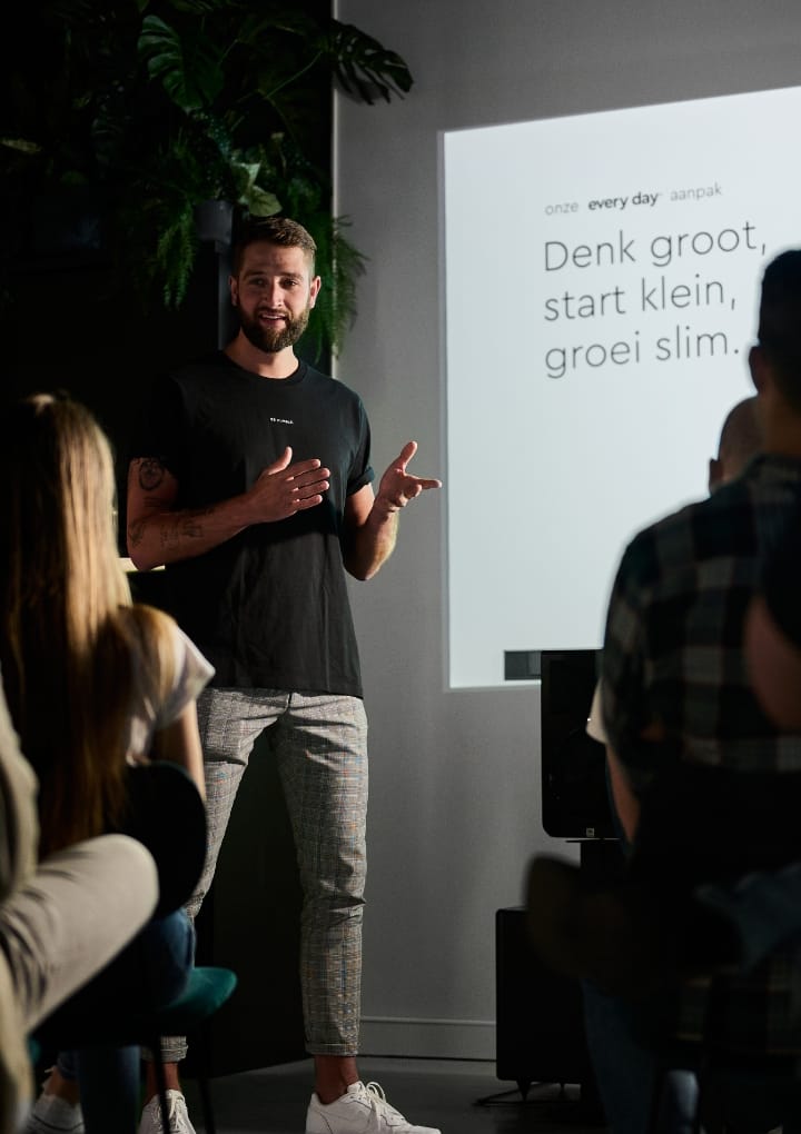 Een presentator staat voor een publiek, achter hem staat een scherm met Nederlandse tekst.