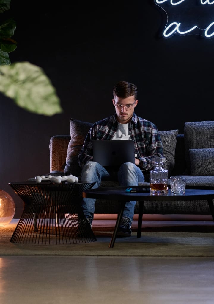 Man op de bank met laptop, zwarte muur, neonreclame op de achtergrond.