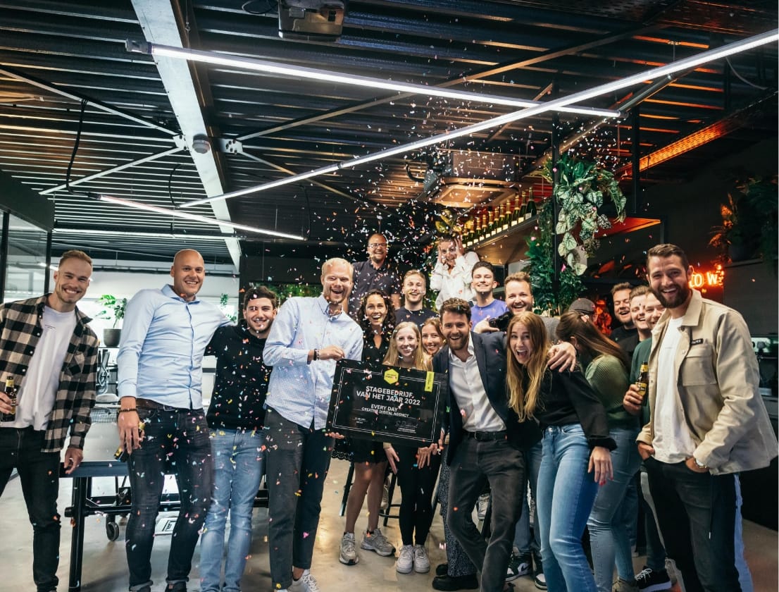 Een vrolijke groep binnen, lachend naar de camera, terwijl er confetti naar beneden vliegt.