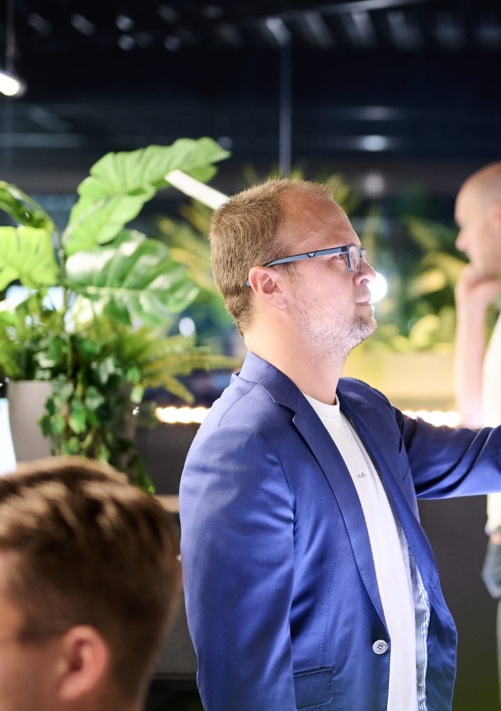 Een persoon met een bril in een blauw jasje schrijft op een bord, omringd door planten.