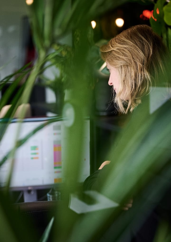 Blondine typt op de computer, gedeeltelijk verborgen door groene bladeren.