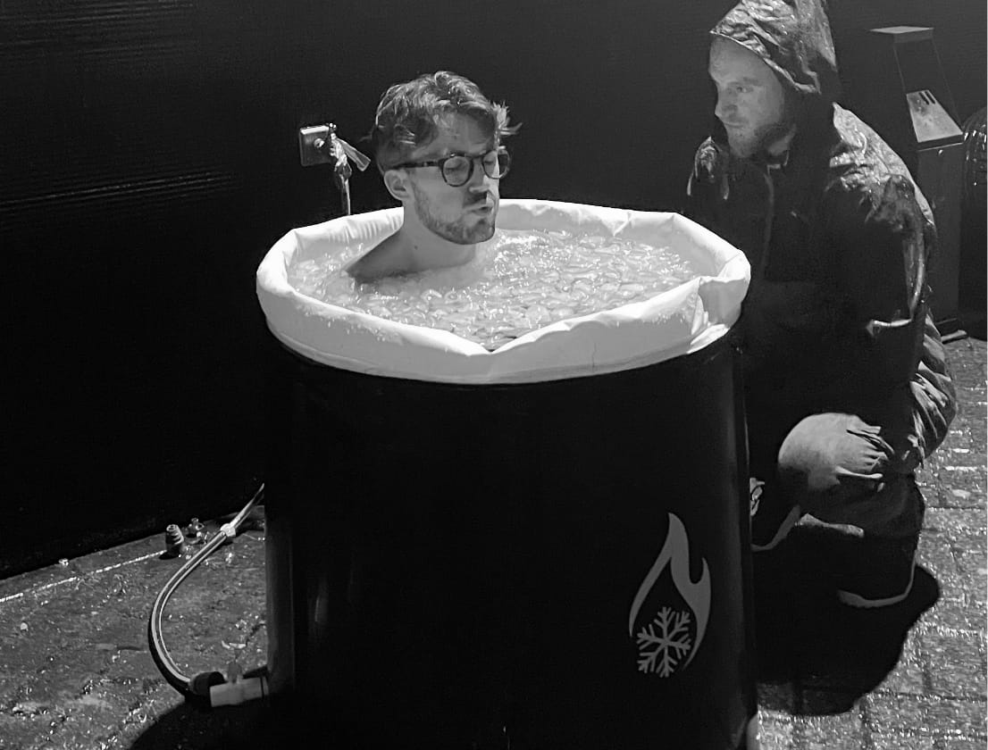 Iemand met een bril op die in een ijsbad ligt, wordt door iemand in een regenjas gadegeslagen.