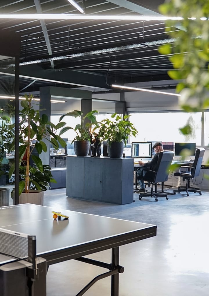 Strak kantoor met veel groen, werkplekken en een pingpongtafel vooraan.