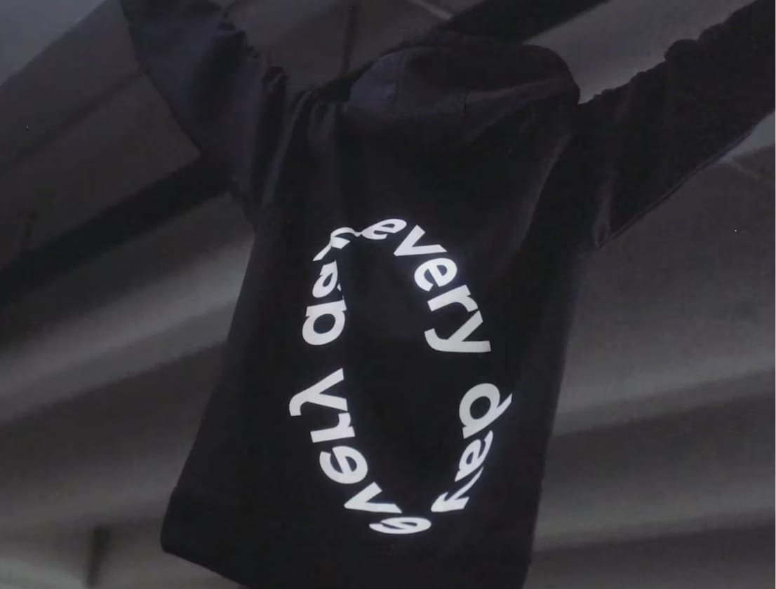 Een persoon in een zwarte hoodie met op de achterkant de tekst 'every day' in cirkelvorm, houdt zijn armen omhoog.