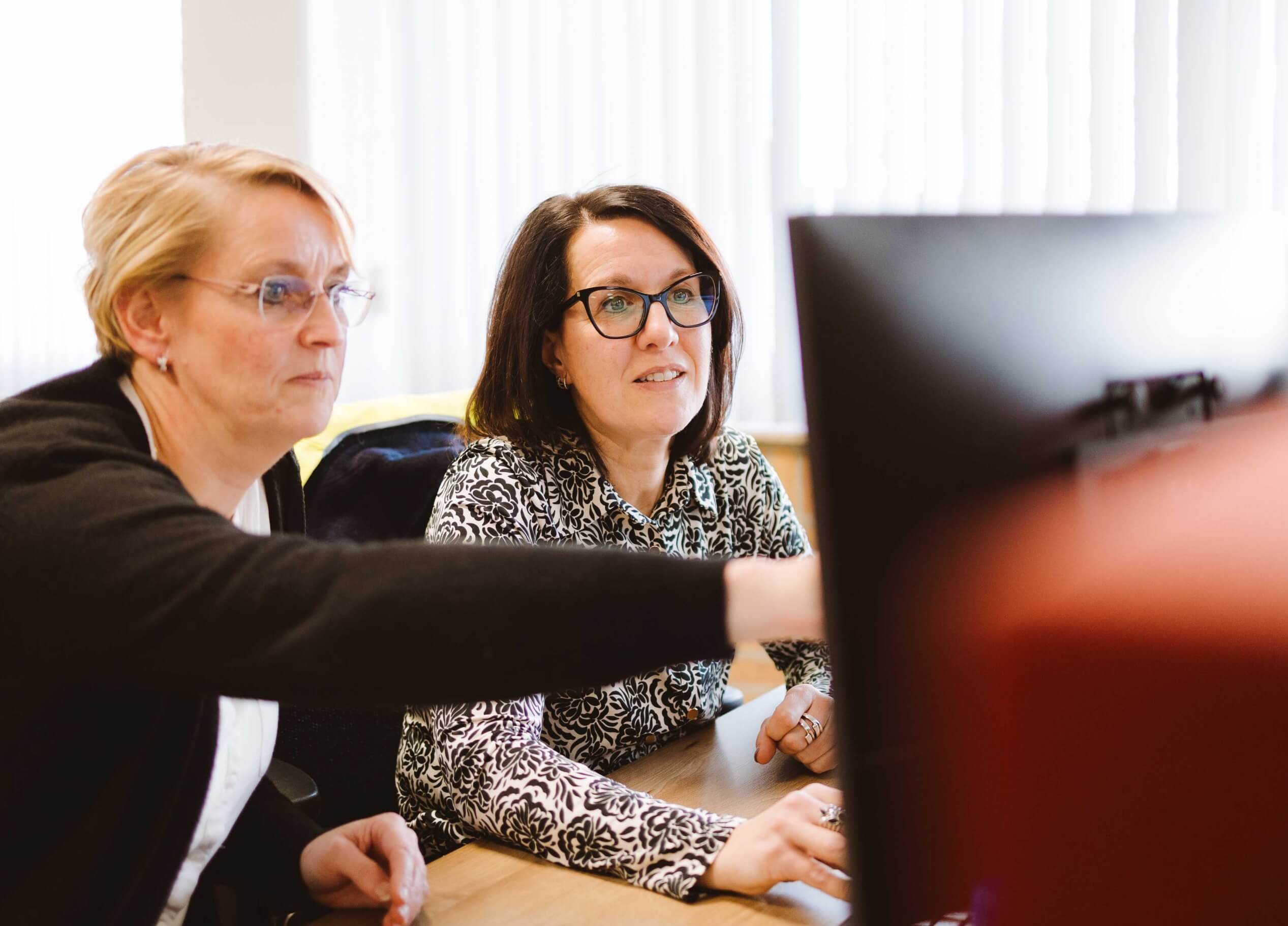 Twee geconcentreerde vrouwen zitten aan een bureau, waarvan er één naar het computerscherm wijst.