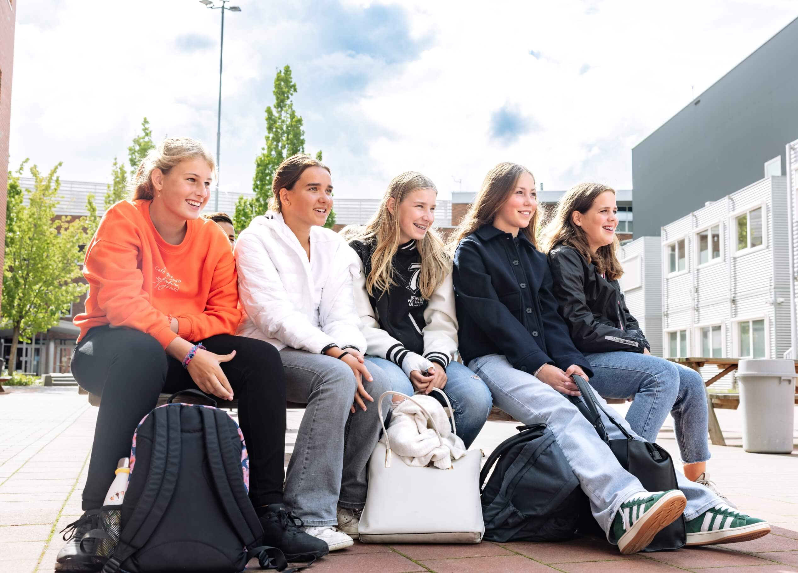 Vijf mensen zitten lachend op een bankje, omringd door hun rugzakken.