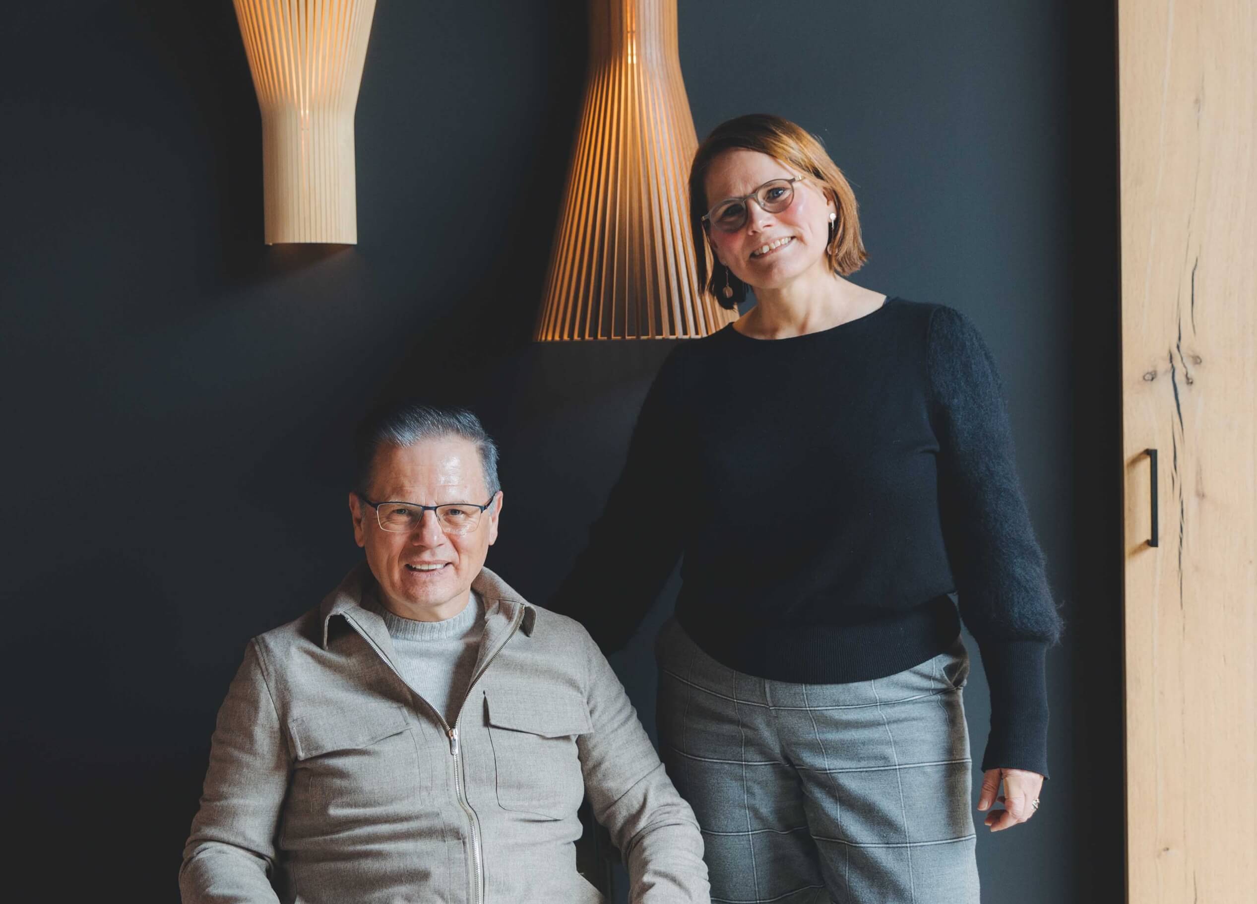 Lachend duo, één zittend en één staand, in een chique kamer verlicht door moderne TunnelmaDesign-lampen op donkere muren.