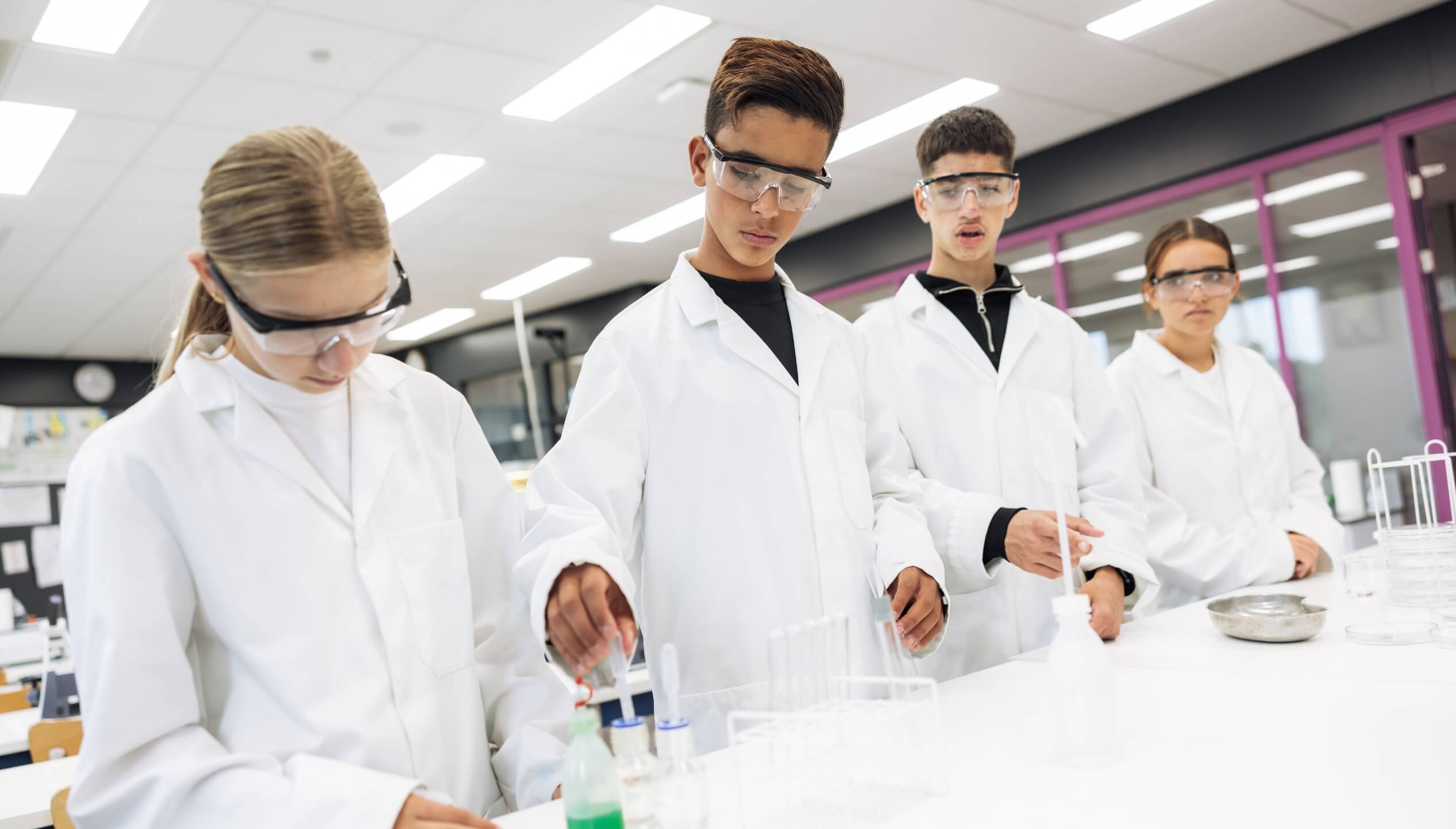 Vier studenten in laboratoriumjassen en met bril op experimenteren met reageerbuisjes op een laboratoriumtafel.