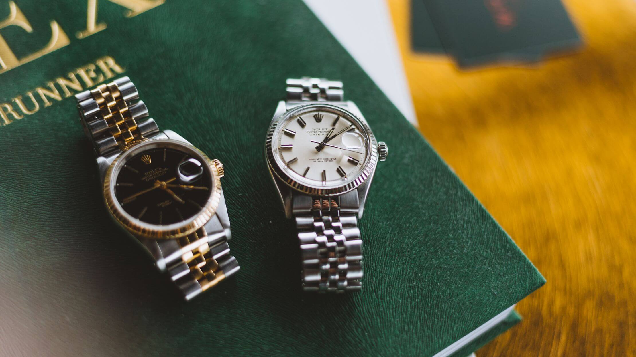 Op een groen boek liggen twee luxe metalen horloges: één met een zwarte wijzerplaat, de ander met een witte.