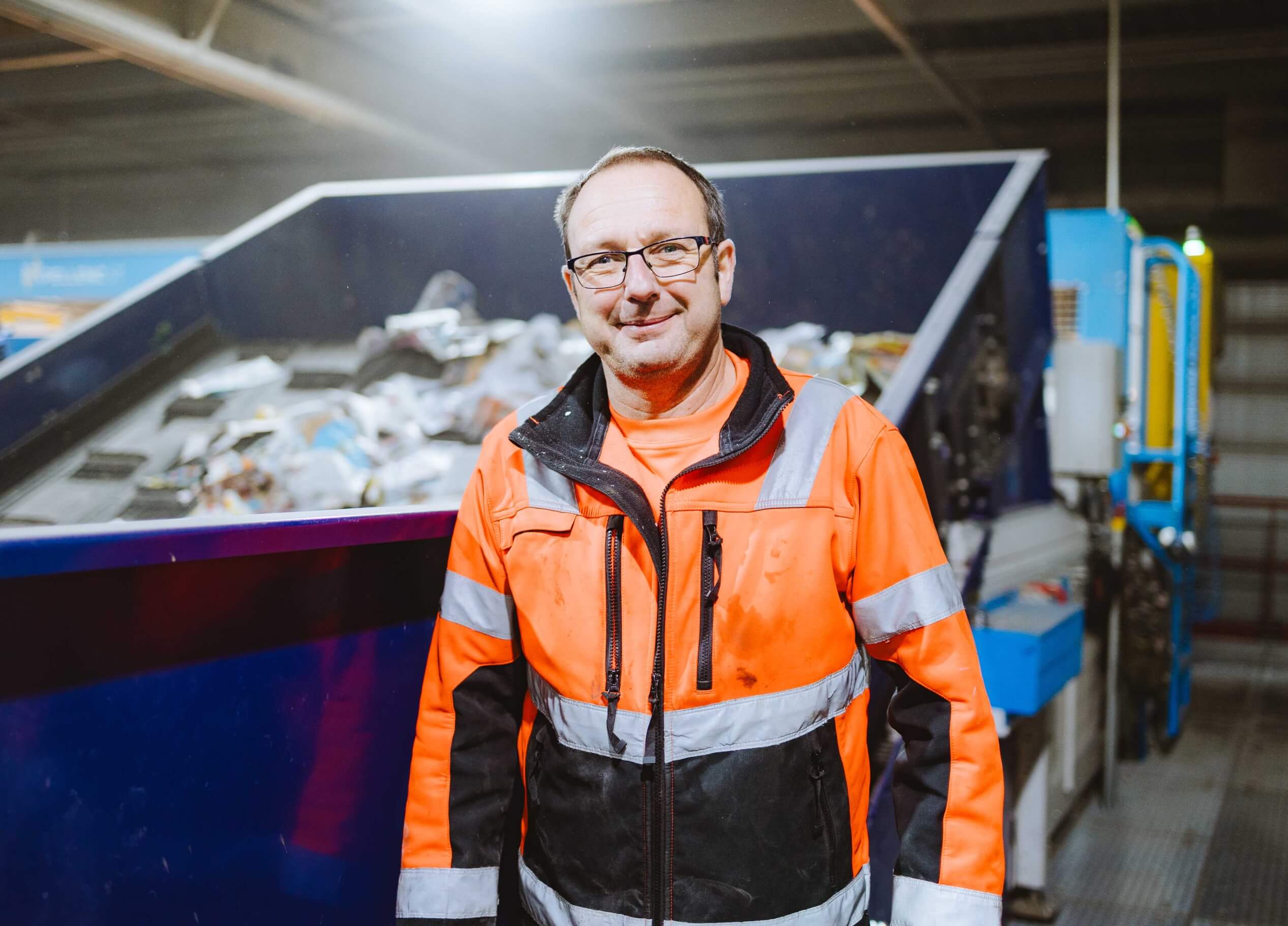 Een man in een oranje veiligheidsvest staat bij een recyclingmachine met daarin allerlei materialen.